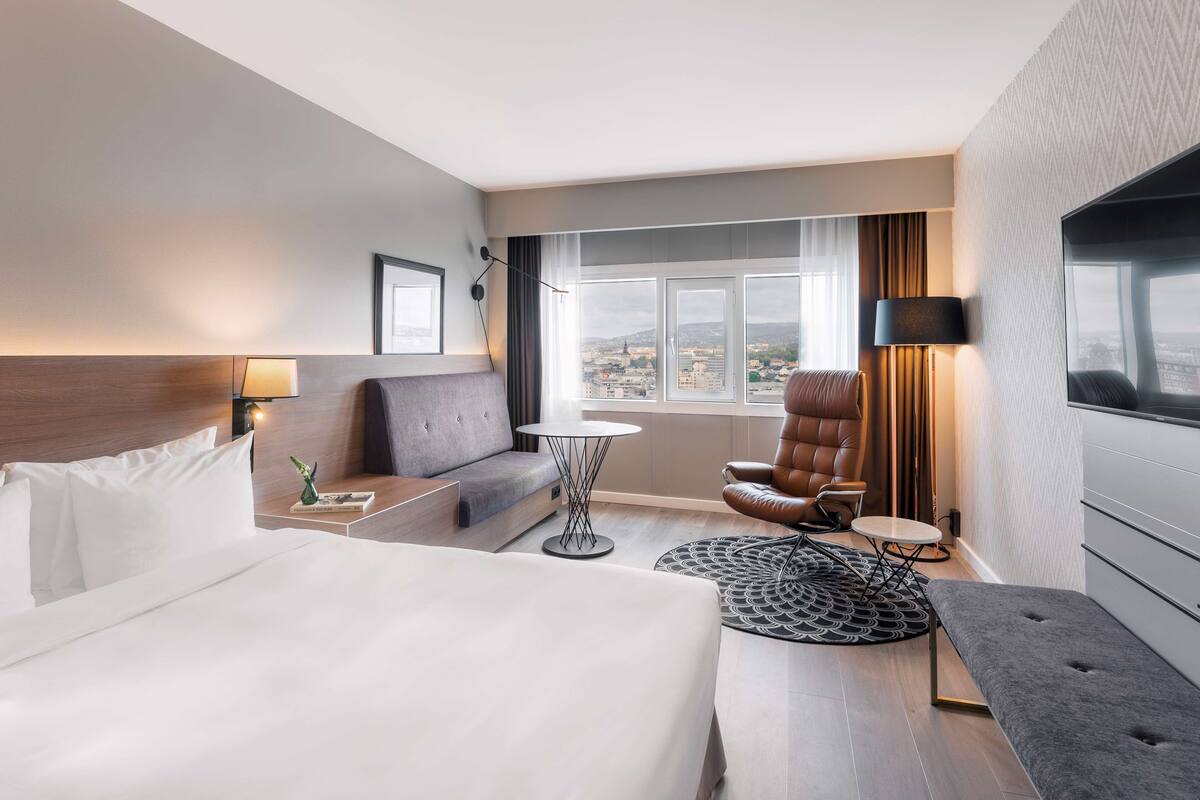 A modern hotel room with a large bed, a sofa, a chair, a small table, and a TV.