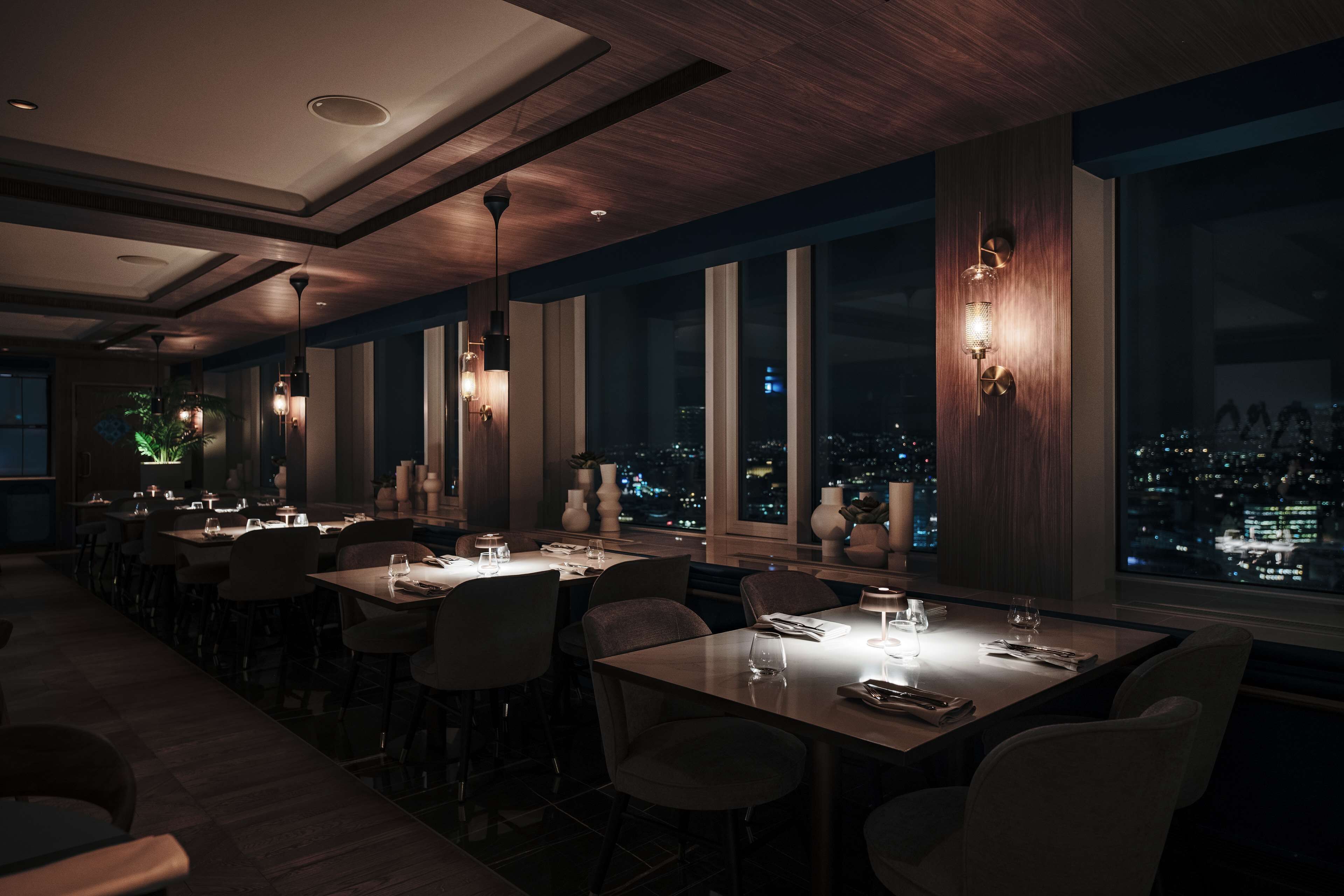 A dimly lit restaurant with multiple tables and chairs, large windows with a city view, and modern pendant lighting.