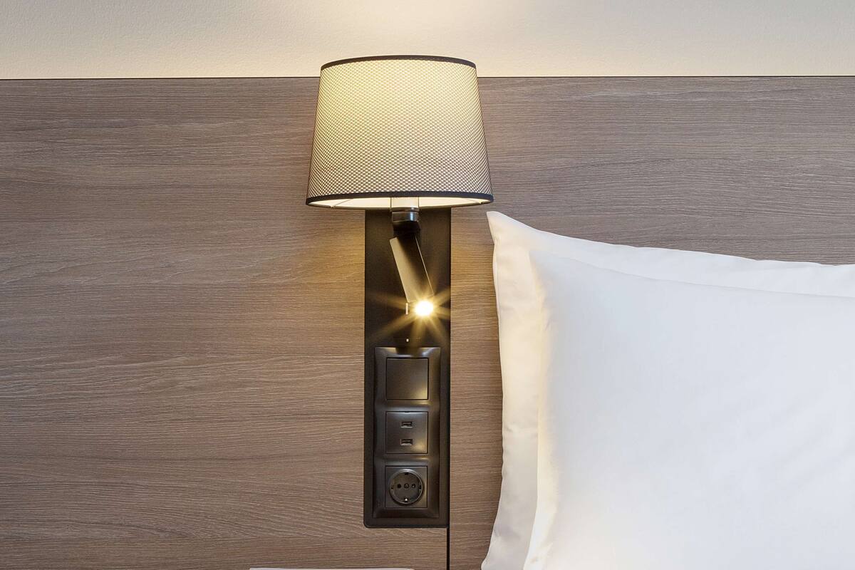 A neatly made bed with a white pillow and a lamp with a mesh shade on a wooden headboard.