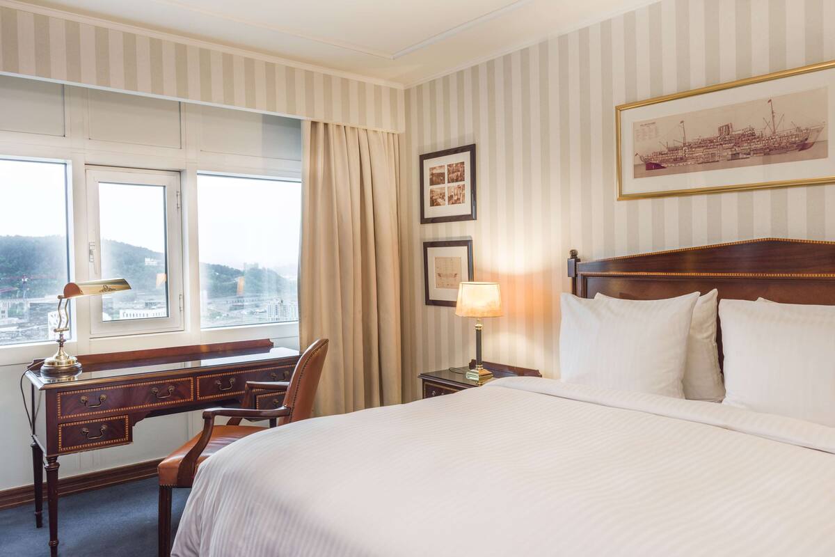 A hotel room with a large bed, a desk, a chair, a lamp, and a window with a view of a cityscape.