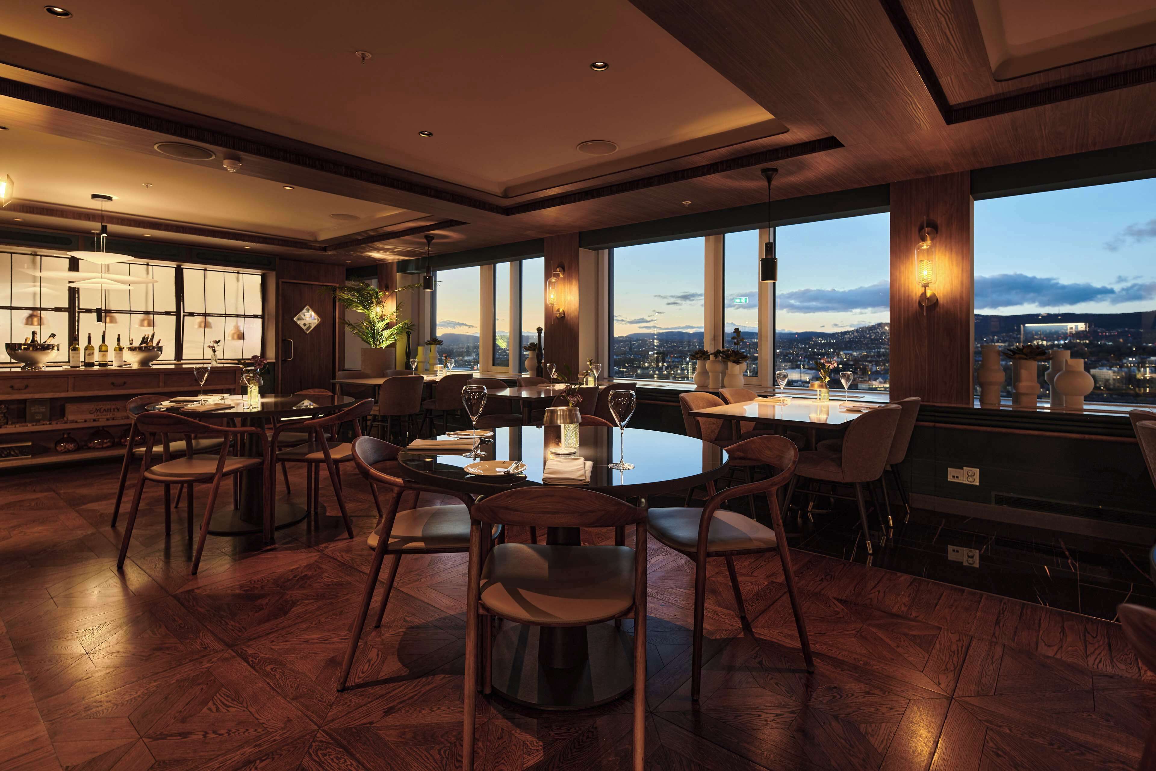 A modern restaurant with a large dining area, multiple tables, and chairs. There are large windows offering a view of the cityscape.