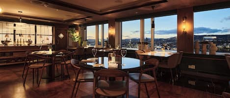 Un restaurante moderno con un amplio salón comedor, varias mesas y sillas. Grandes ventanales ofrecen vistas panorámicas de la ciudad.