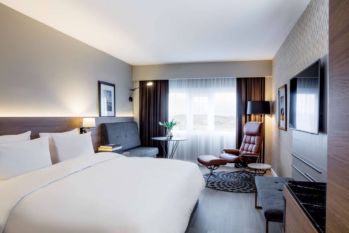 A hotel room with a large bed, a desk, a chair, a small table, and a TV.