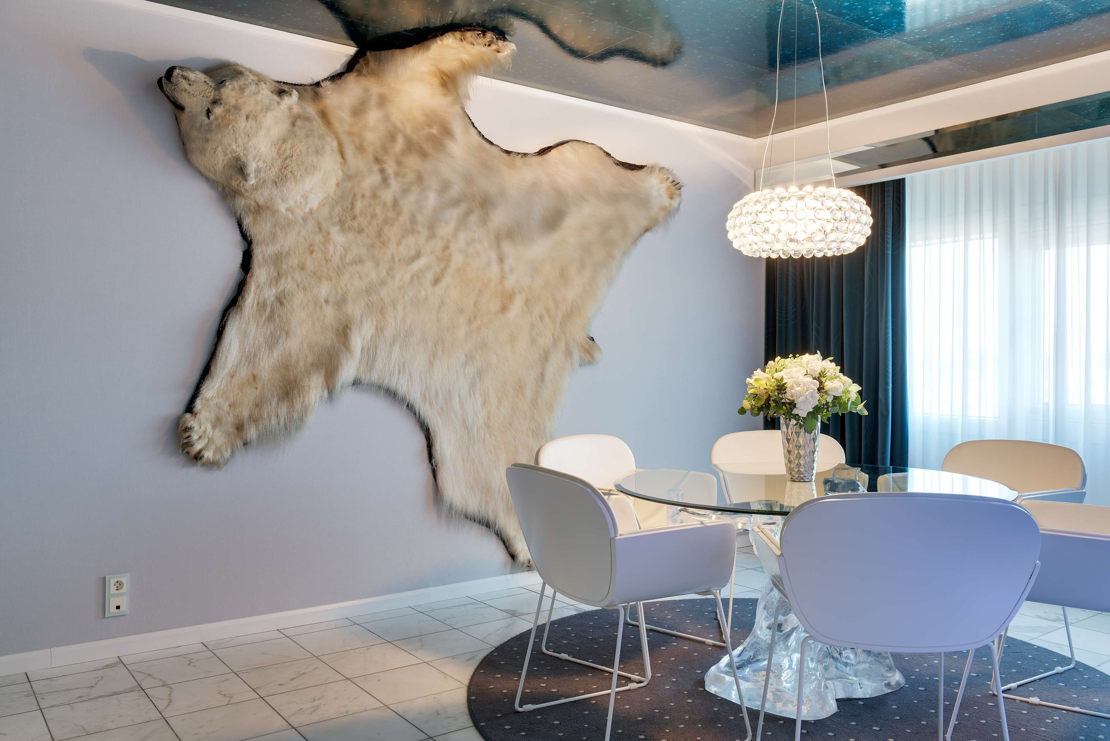 Een moderne eetruimte met een glazen tafel, witte stoelen en een opgehangen dierenhuid aan de muur.