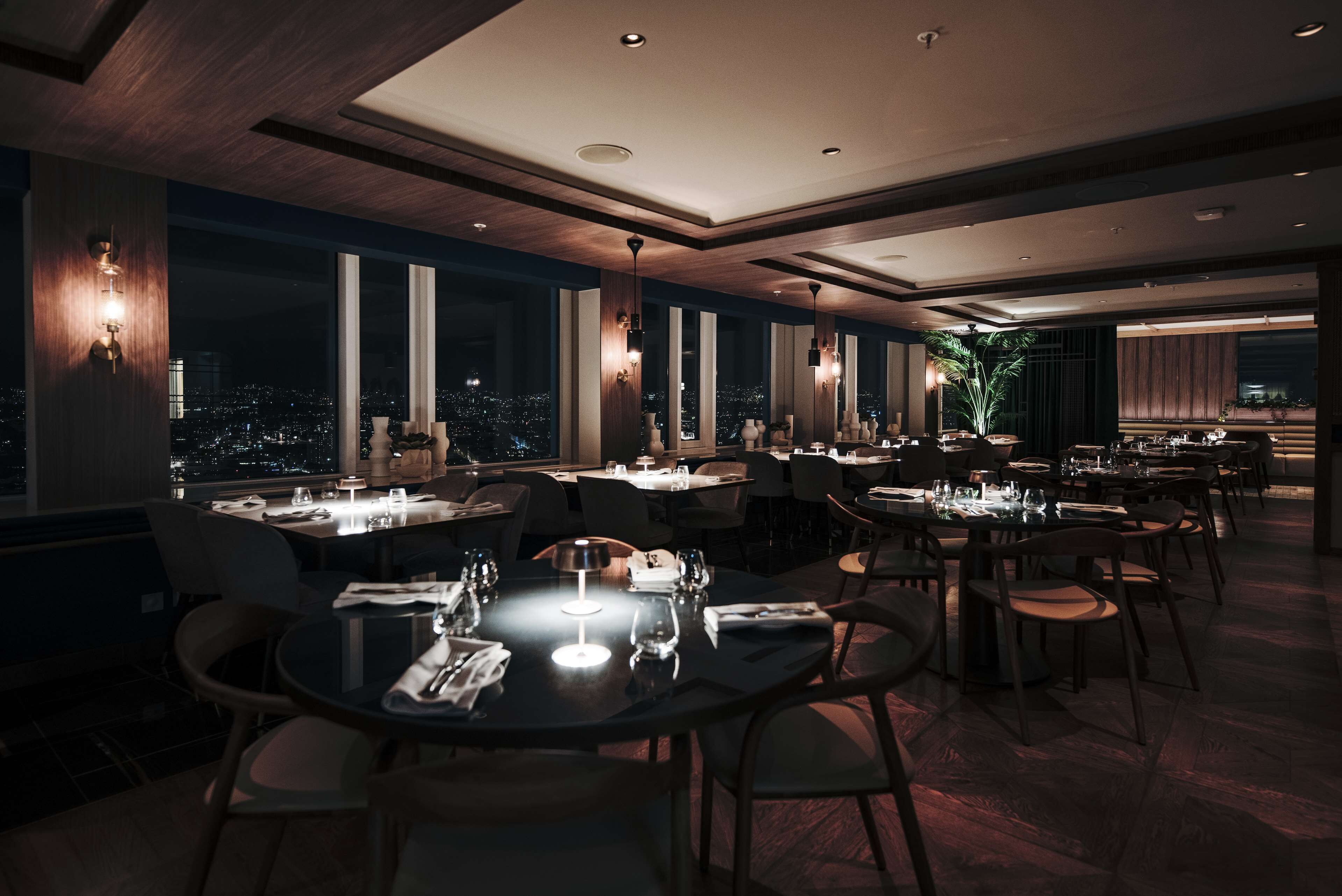 Een modern restaurant met ronde tafels en stoelen, grote ramen met uitzicht op de stad en warme verlichting.