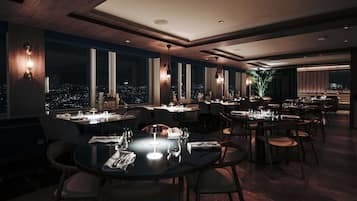 Een modern restaurant met ronde tafels en stoelen, grote ramen met uitzicht op de stad en warme verlichting.