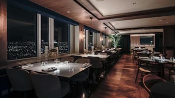 A modern dining area with long tables, chairs, and large windows offering a city view.