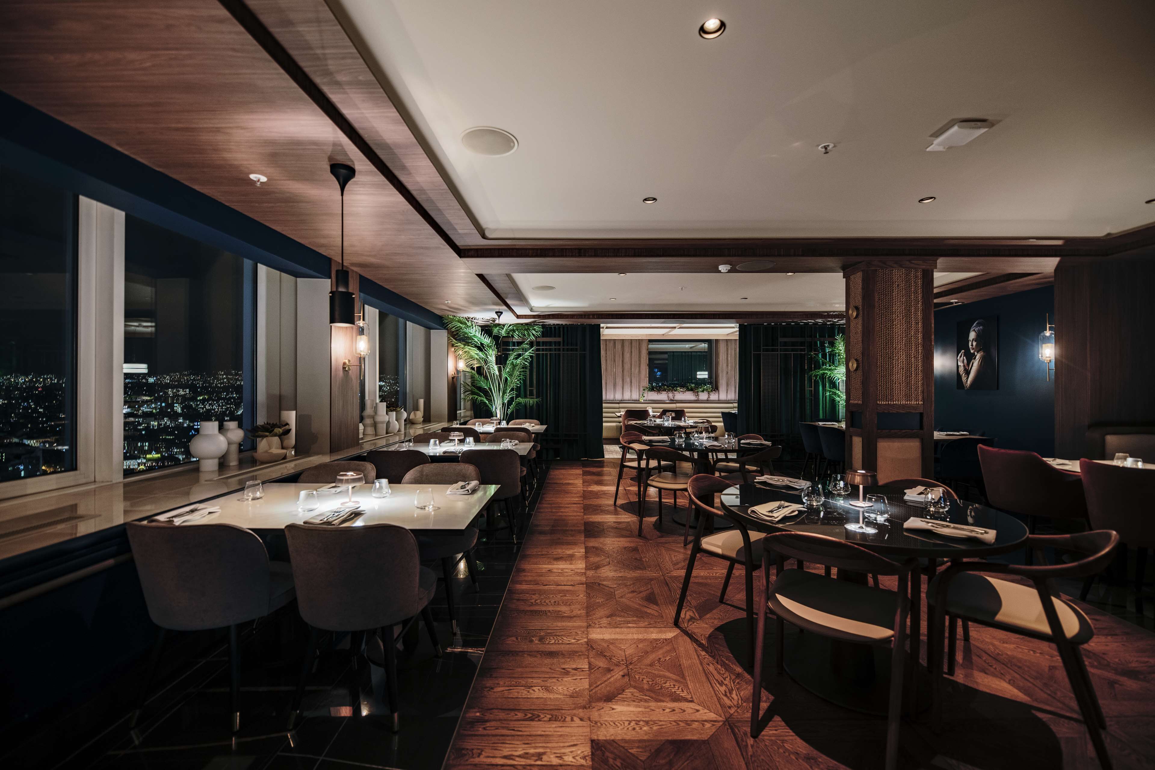 A modern restaurant with a long dining area, large windows, and a city view.