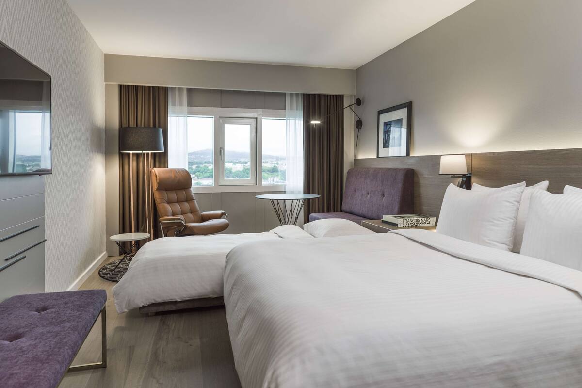 A hotel room with two beds, a chair, a small table, a lamp, and a view of the city.