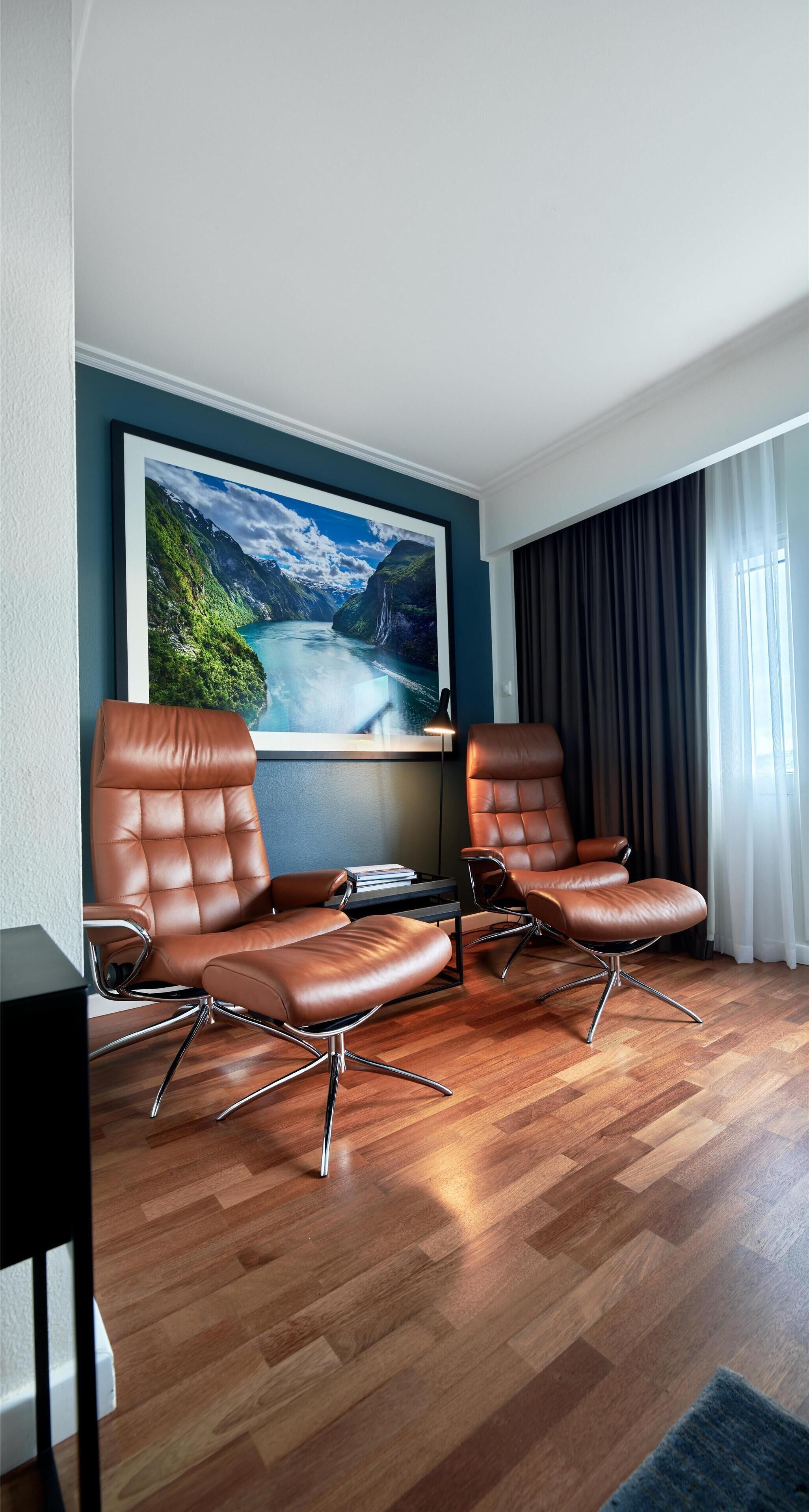 A modern hotel room with a large window, two leather armchairs, a wooden floor, and a framed landscape painting on the wall.