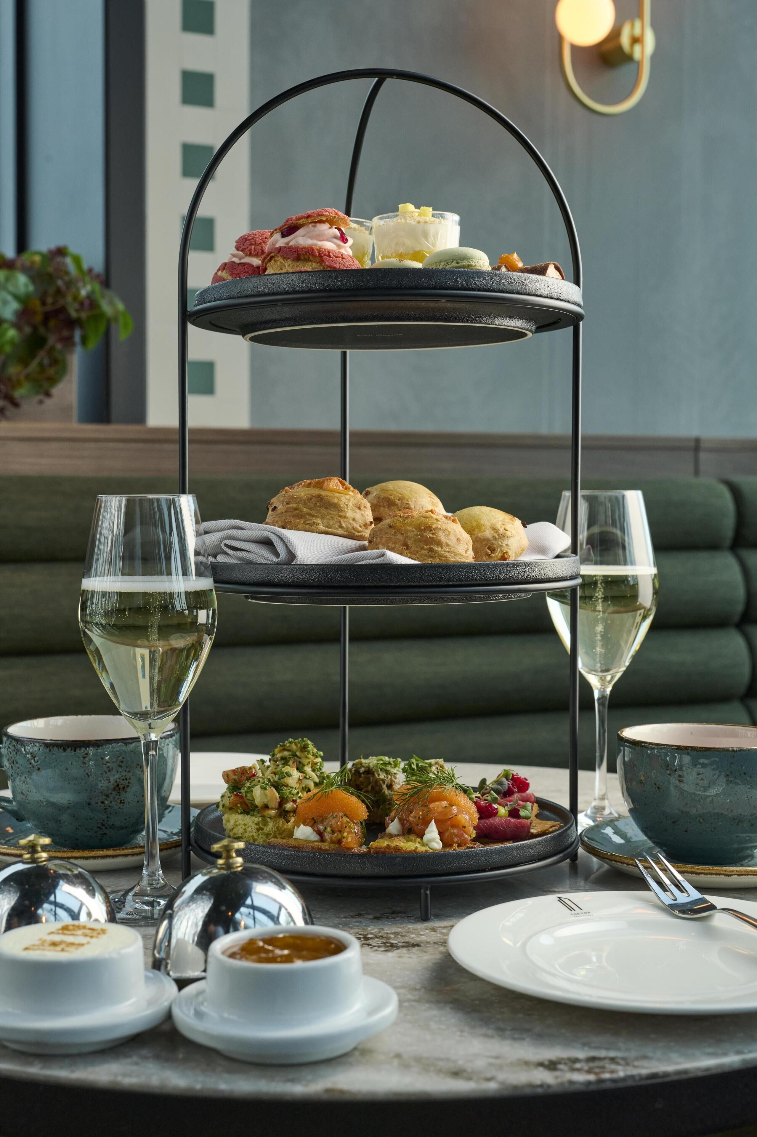 A three-tiered dessert stand with pastries and a plate of food, accompanied by two glasses of white wine and a green upholstered sofa in the background.