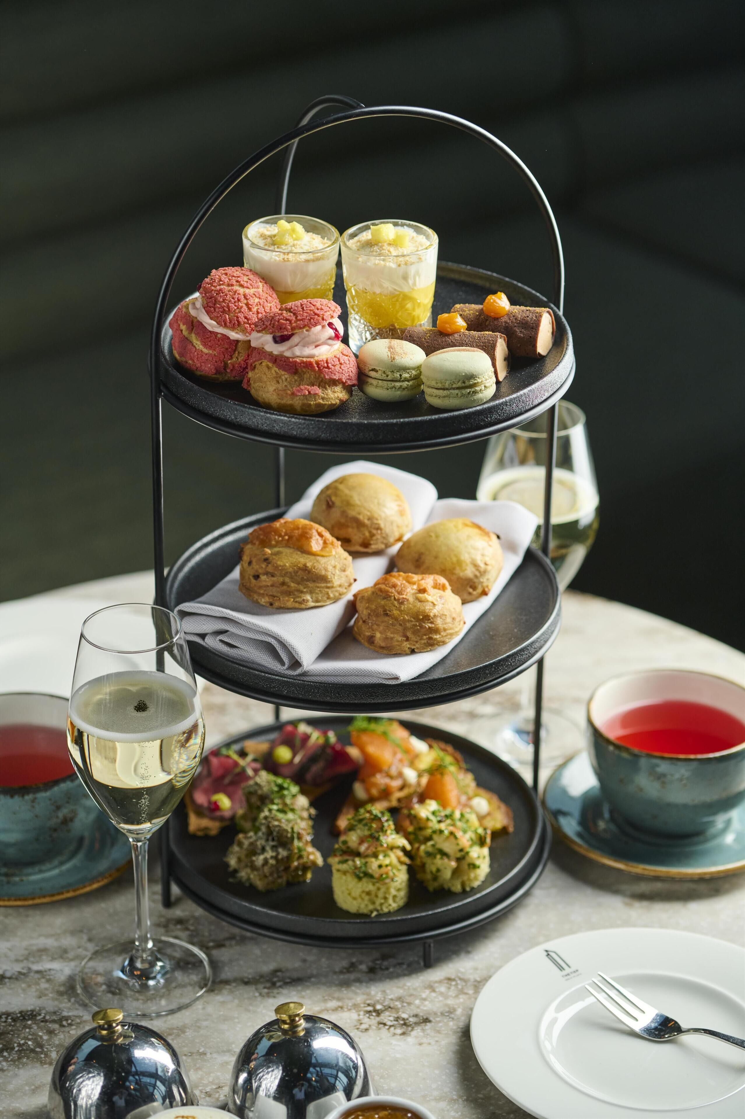 A three-tiered dessert stand with macarons, scones, and a variety of desserts, accompanied by glasses of white wine and tea.