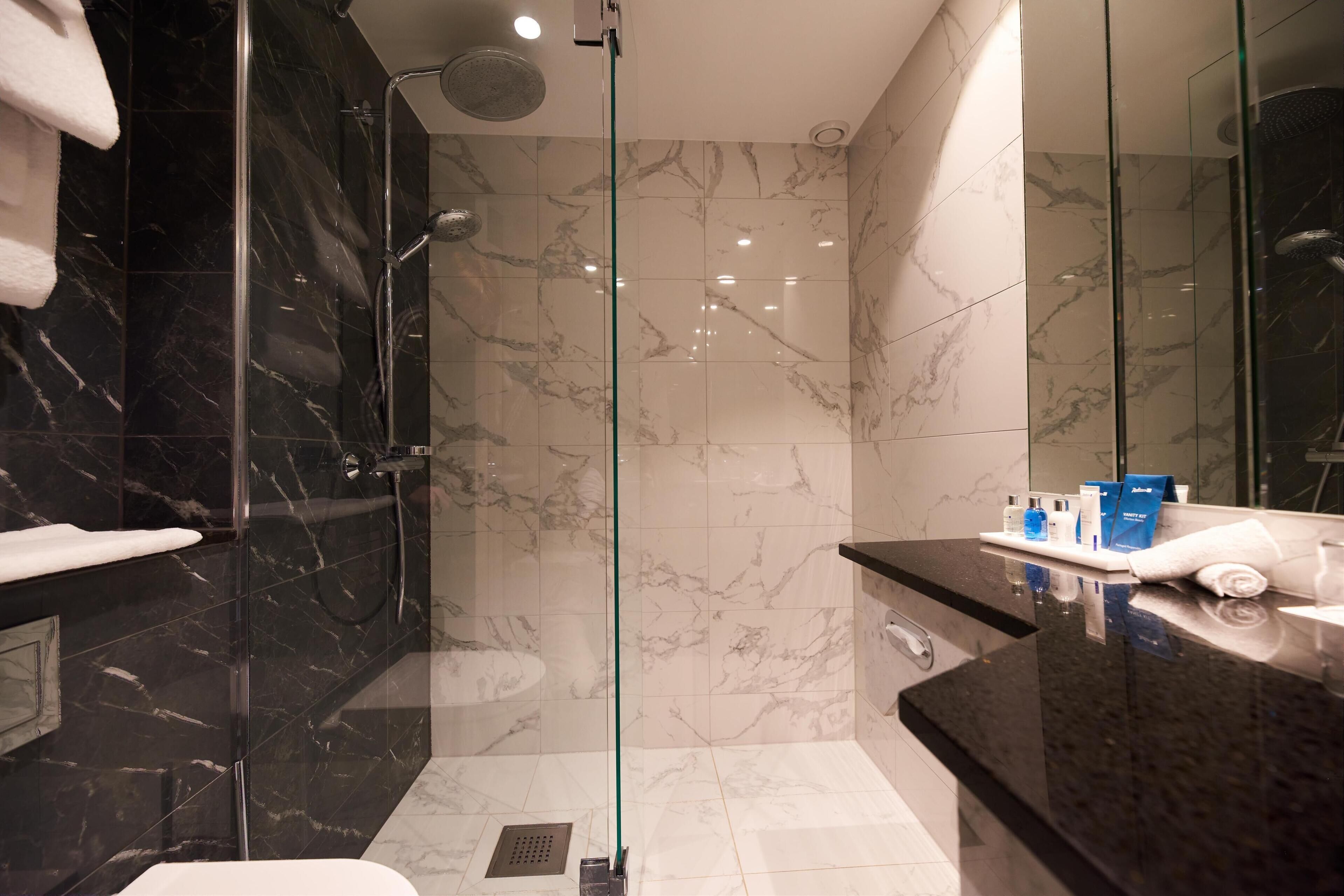 Un baño moderno con ducha de vidrio, azulejos de mármol y tocador con artículos de aseo.