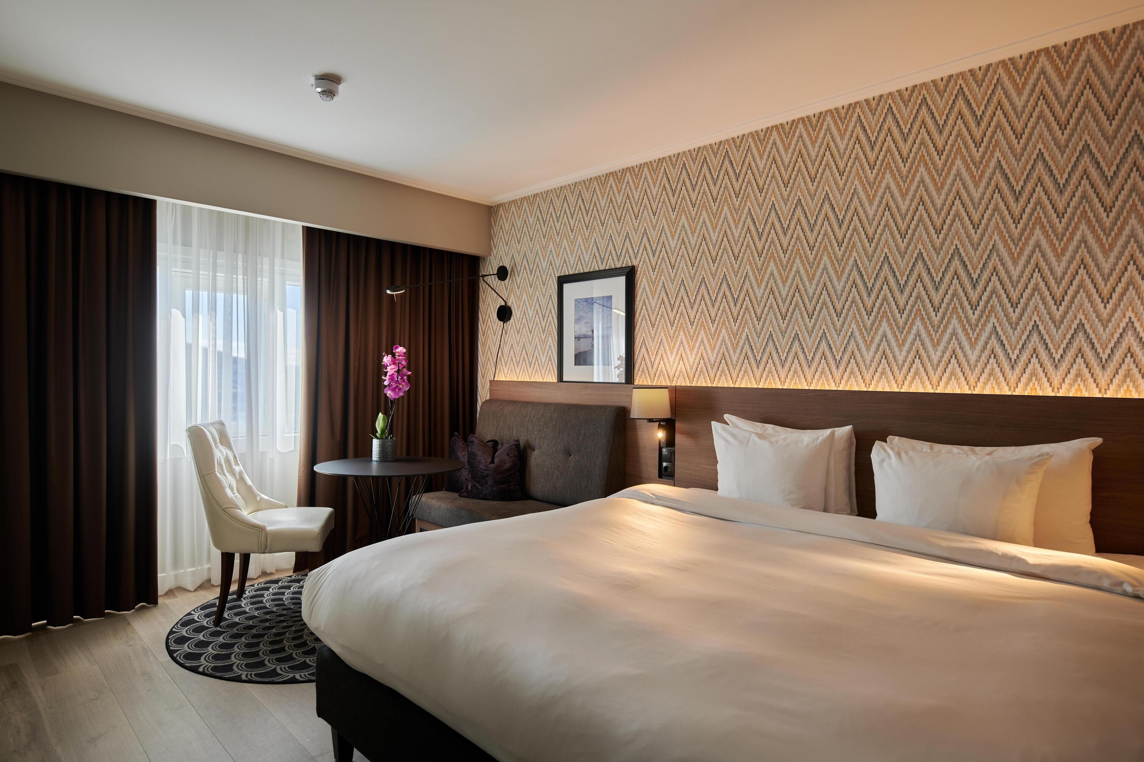 A hotel room with a large bed, a desk, a chair, and a small table with a vase of flowers.