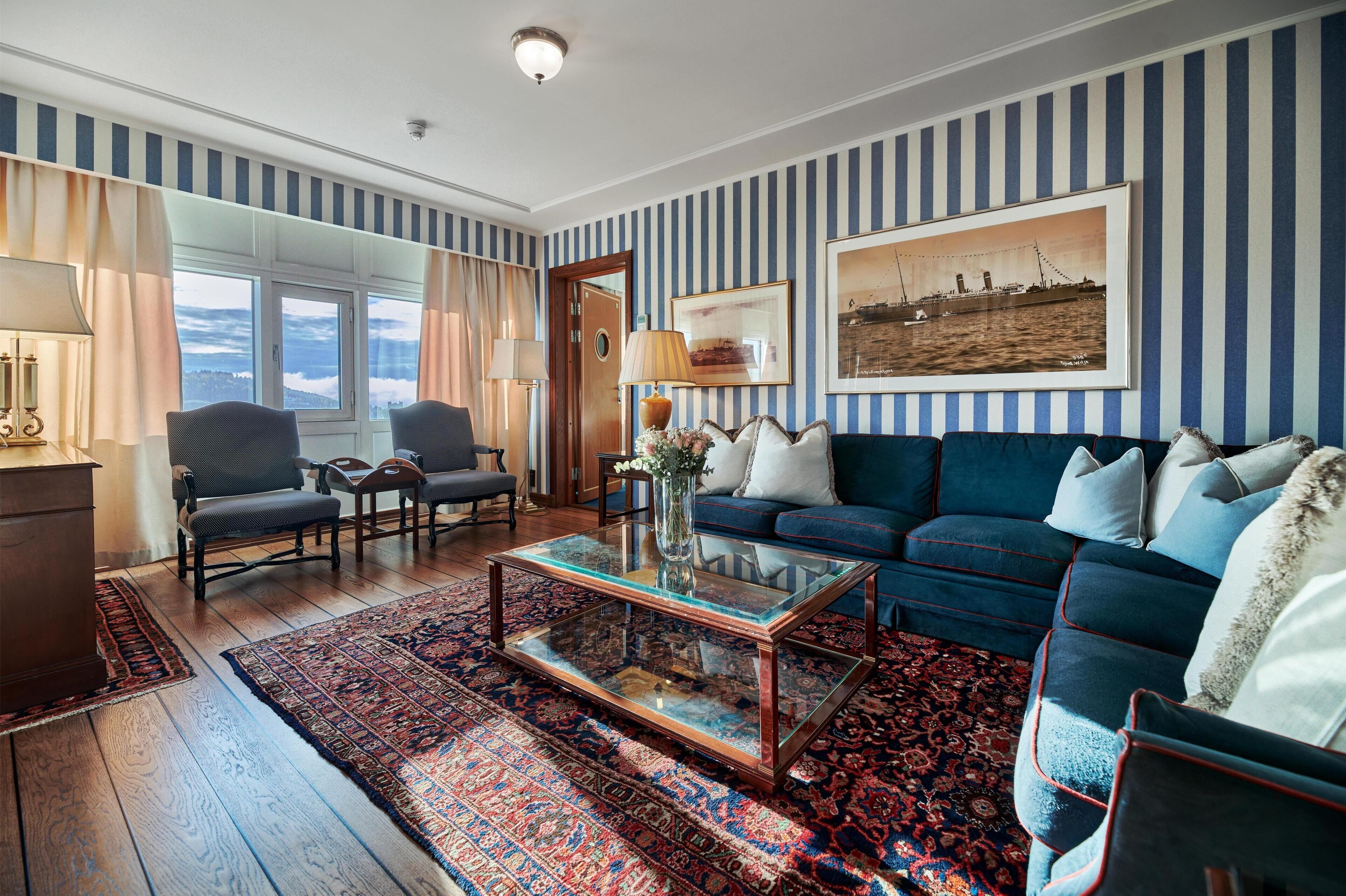 A room with a blue-striped wall, a sofa, a glass coffee table, and a patterned rug.
