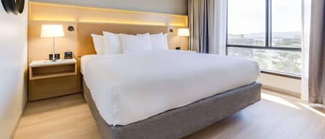 A hotel room with a large bed, bedside lamps, a nightstand, and a view of the city through a large window.
