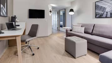 A modern hotel room with a desk, laptop, grey sofa, ottoman, and a flat-screen TV.