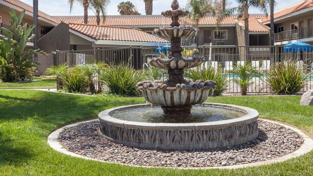 A circular fountain with multiple tiers, surrounded by a gravel border and grass. In the background, there are palm trees and a building with a tiled roof.