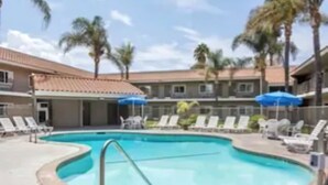 A swimming pool with lounge chairs and umbrellas, surrounded by palm trees and a building with a tiled roof.