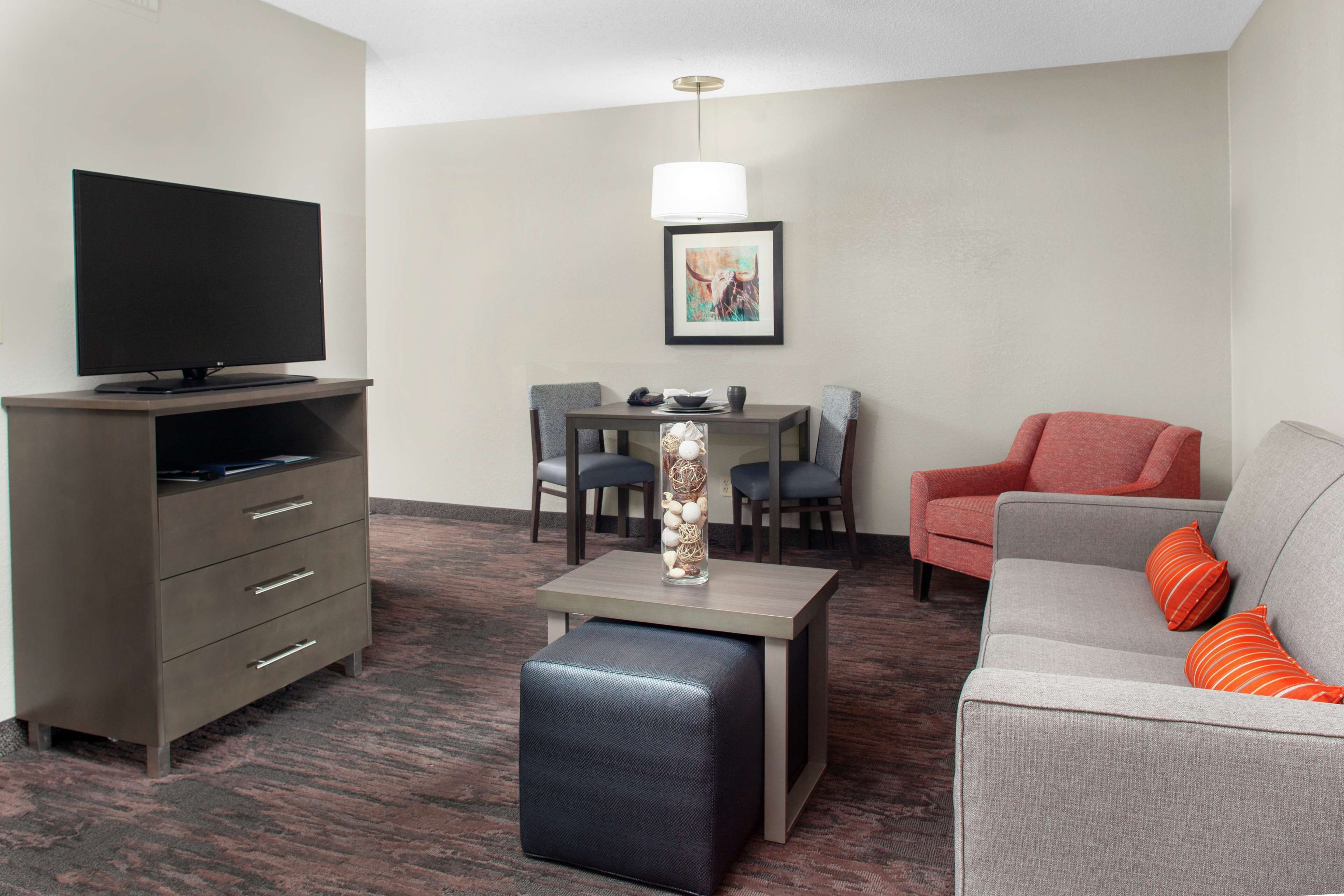 A hotel room with a flat-screen TV, a dining table with chairs, a sofa with cushions, a small ottoman, and a framed picture on the wall.