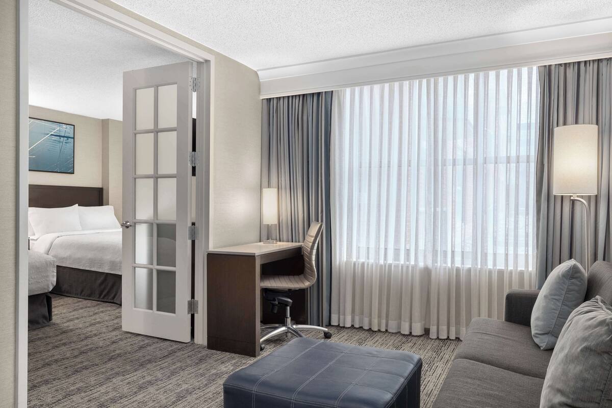 A hotel room with a bed, a desk with a chair, a sofa, and a large window with curtains.