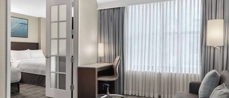 A hotel room with a bed, a desk with a chair, a sofa, and a large window with curtains.