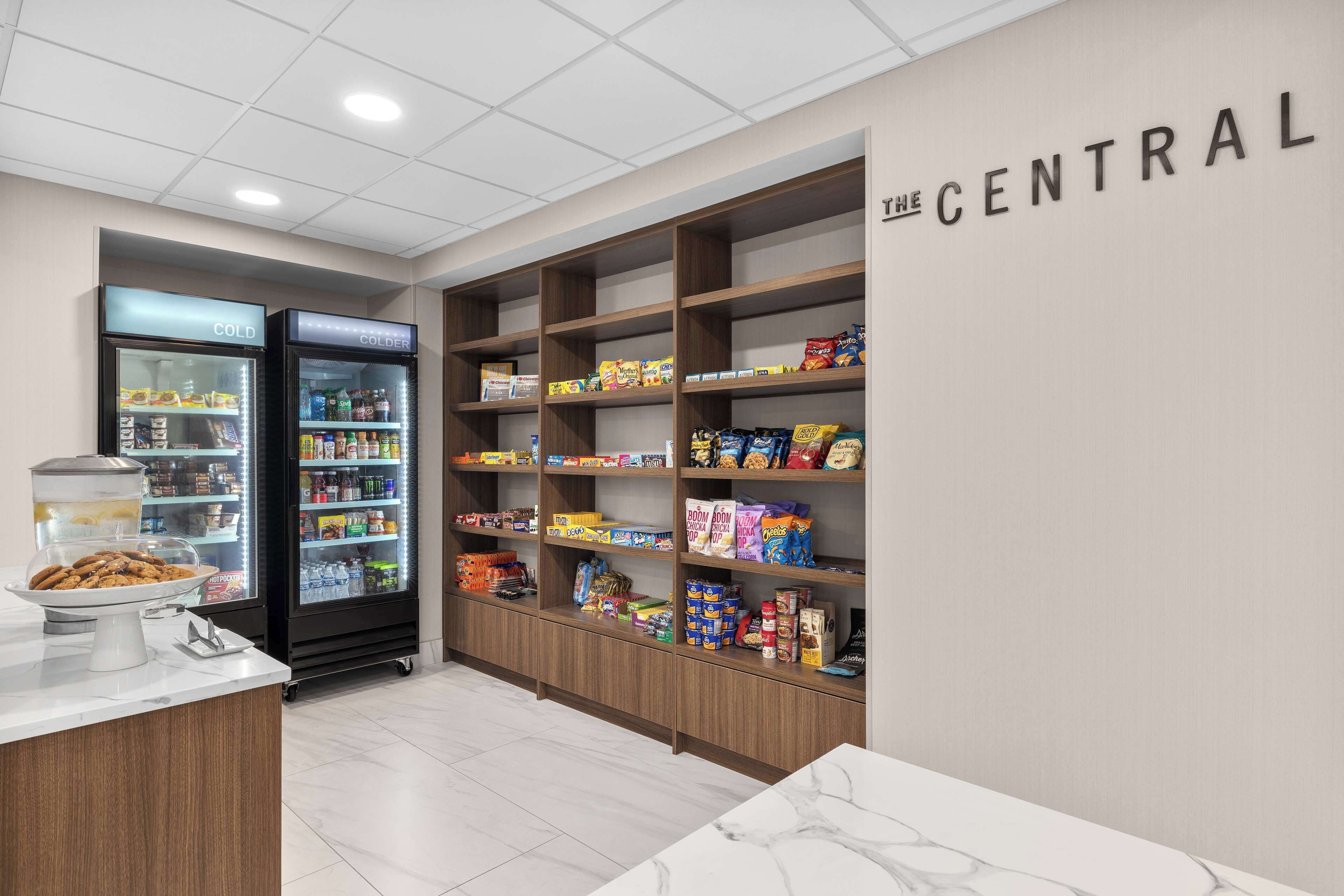 Un área de comidas con una estación de autoservicio, dos refrigeradores con las etiquetas 'FRÍO' y 'ENFRIADOR', y estantes llenos de refrigerios y bebidas.