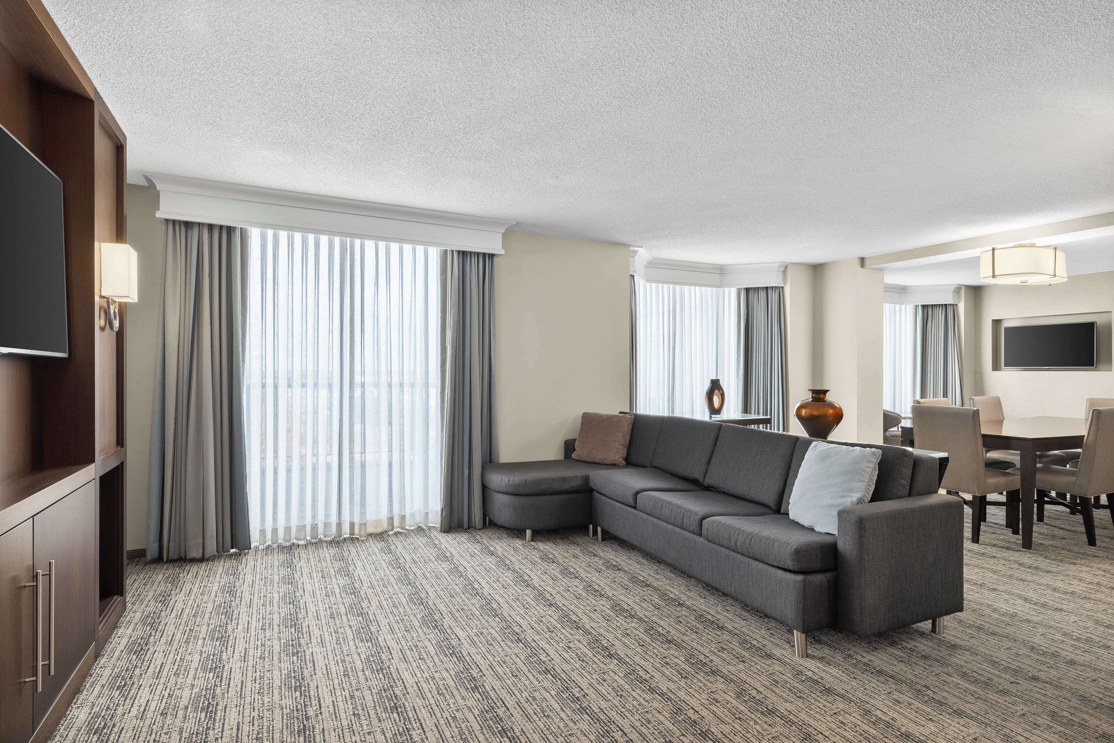 A hotel room with a sectional sofa, a flat-screen TV, a dining area, and large windows with curtains.