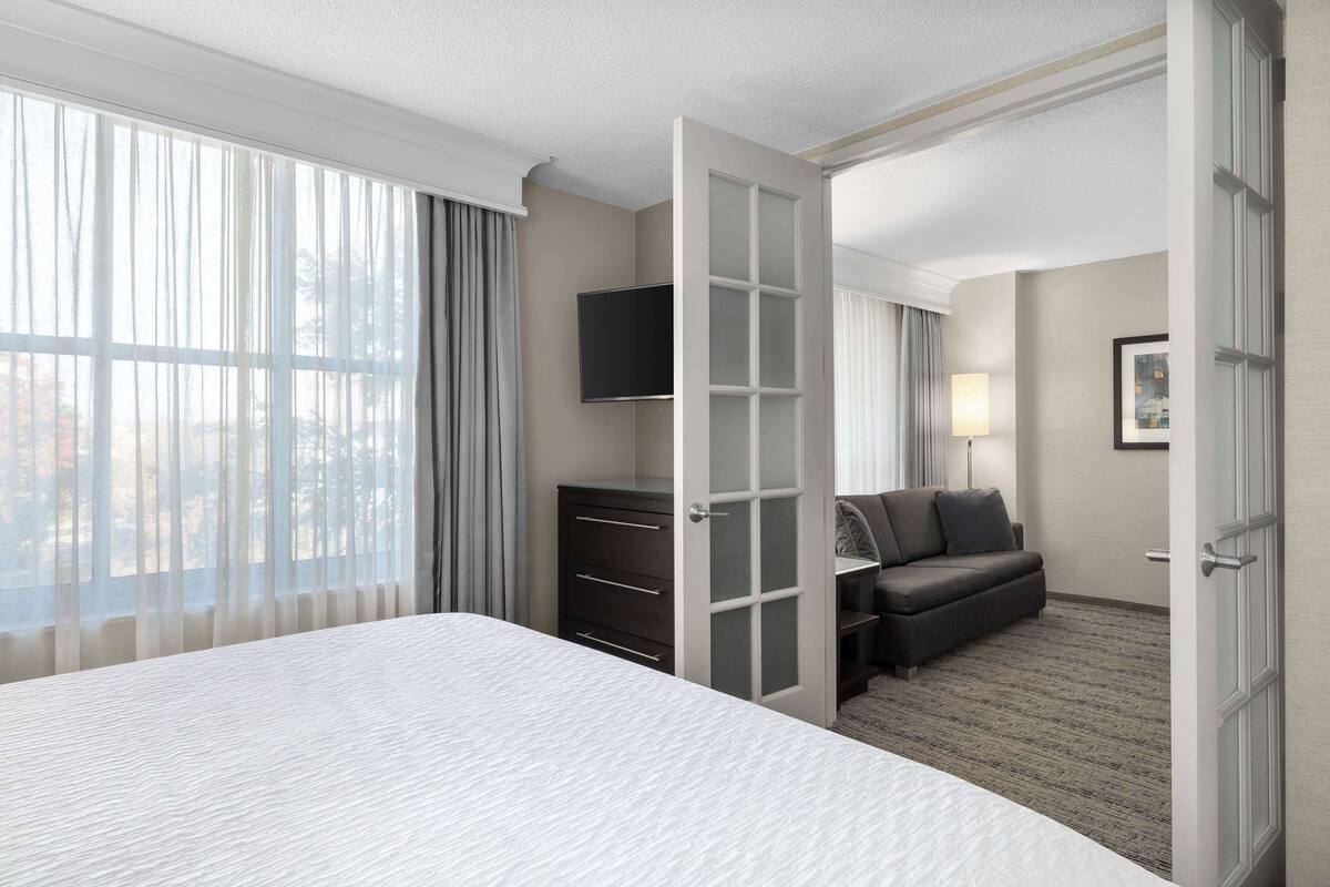 A hotel room with a large bed, a desk with drawers, a flat-screen TV, a sofa, and a window with curtains.
