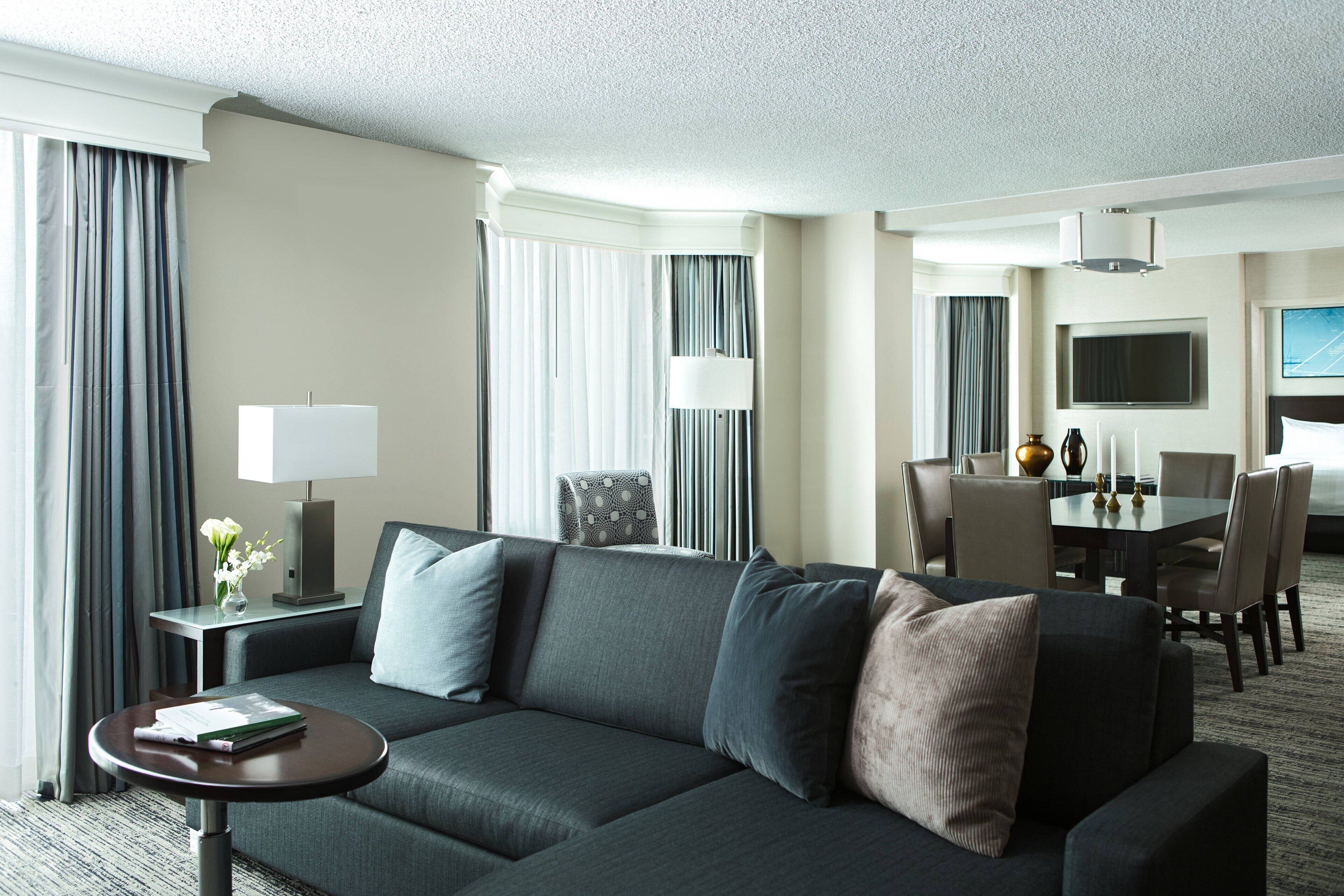 Una habitación de hotel moderna con un sofá gris, una pequeña mesa redonda con una lámpara, un rincón de comedor con una mesa y sillas, y un televisor montado en la pared.