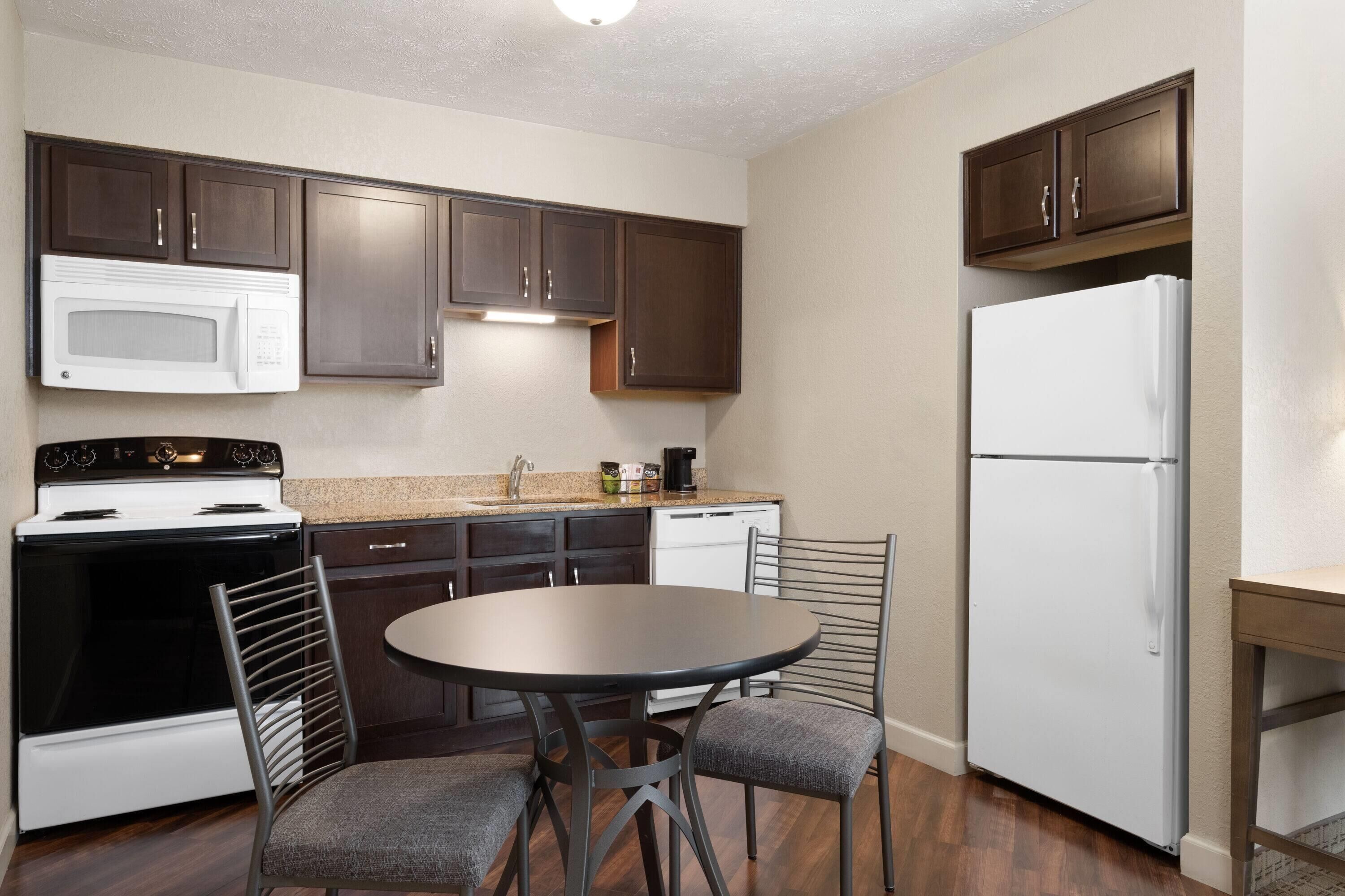 A compact kitchen with dark wooden cabinets, a white microwave, a stove, a dishwasher, and a refrigerator. There is a round table with two chairs.