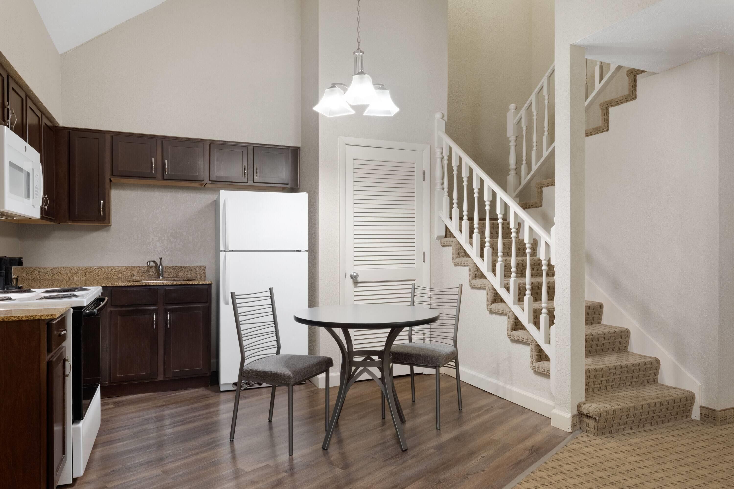 A kitchen with dark wooden cabinets, a white refrigerator, a dining table with two chairs, and a staircase with white railings.