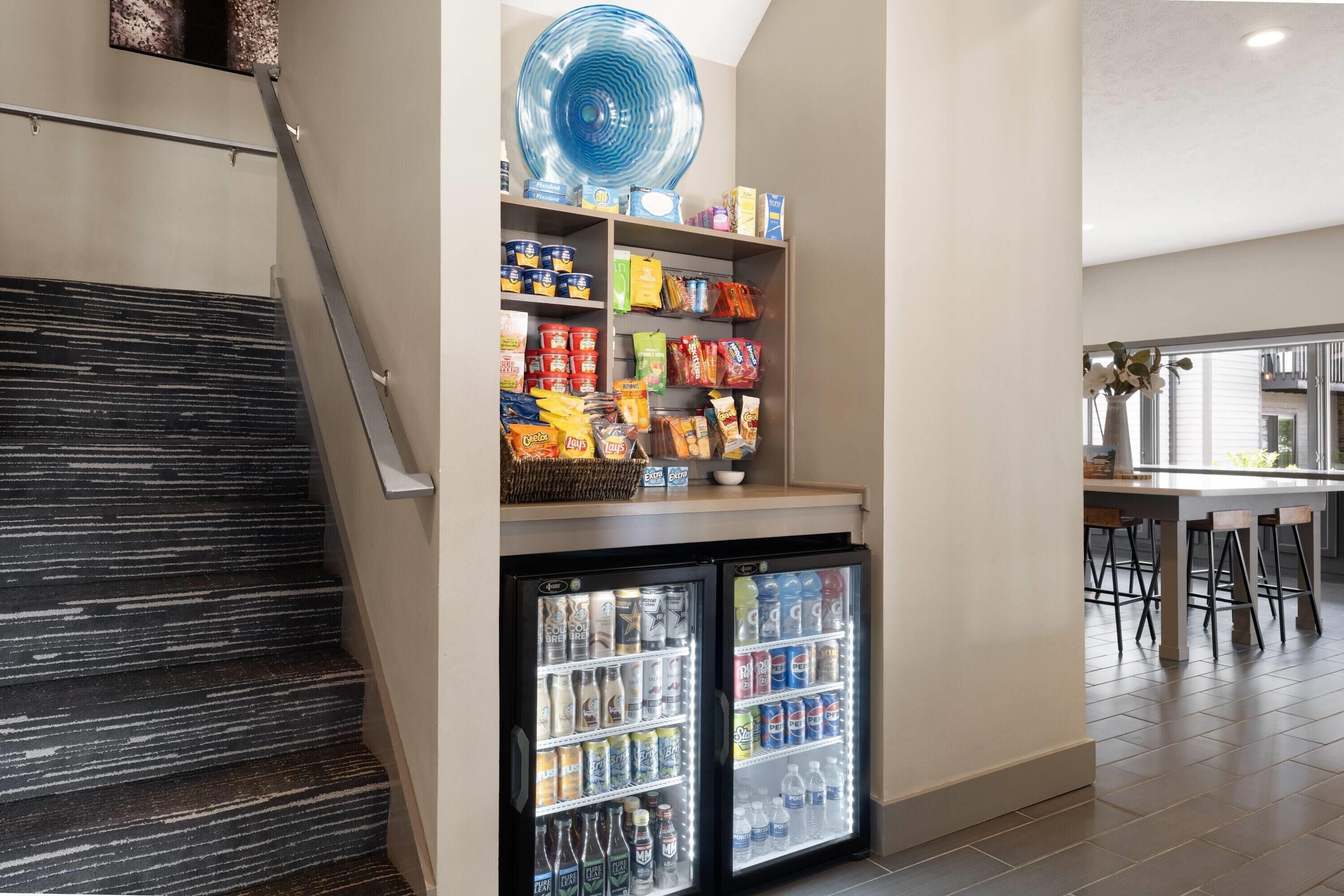 Un coin cuisine équipé d’un réfrigérateur rempli de boissons, d’une étagère avec des en-cas et d’un espace repas avec des tabourets de bar.