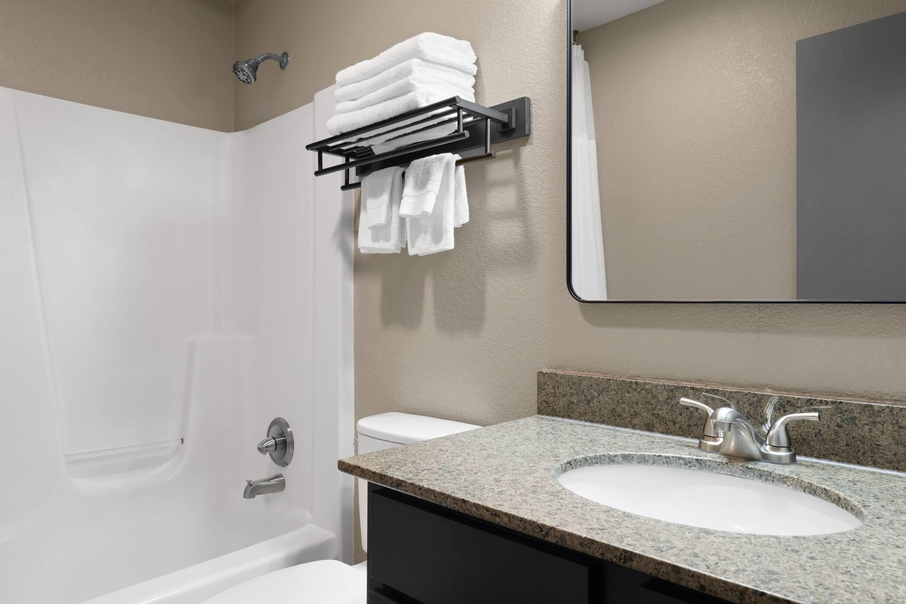 Un baño con una bañera blanca, un lavabo con encimera de granito y un perchero con toallas dobladas.