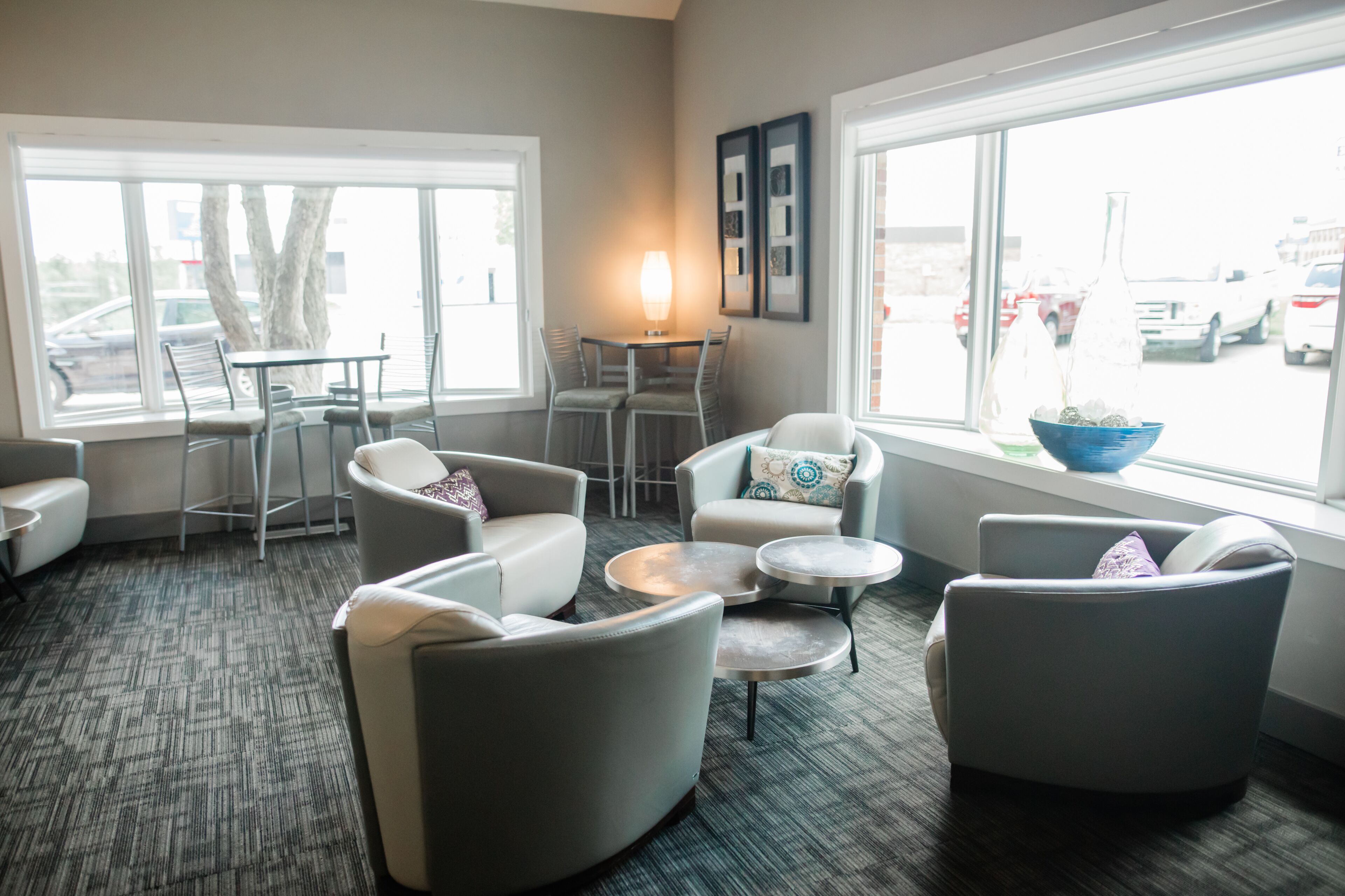A modern lounge area with seating, a table, and a view of the parking lot through large windows.