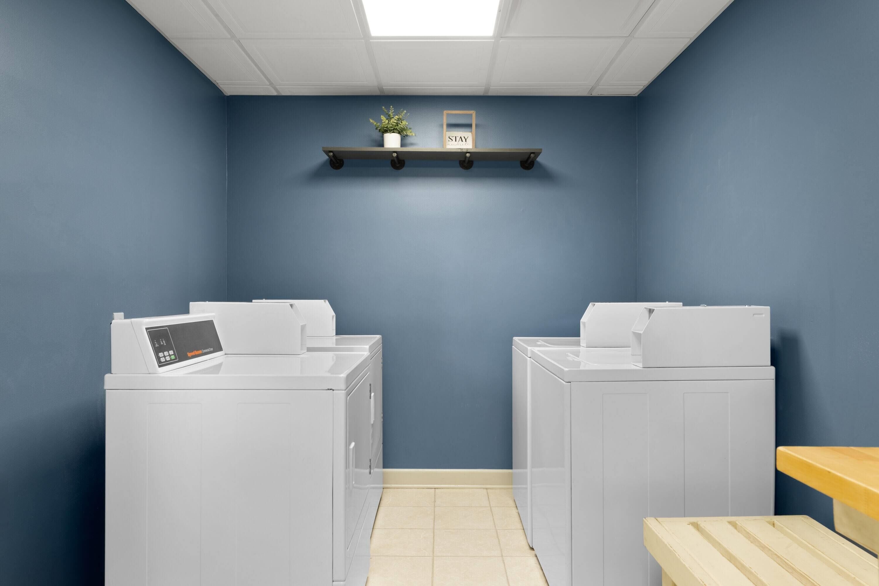 A laundry room with two white washers and dryers, a shelf with a plant and a sign, and a wooden bench.