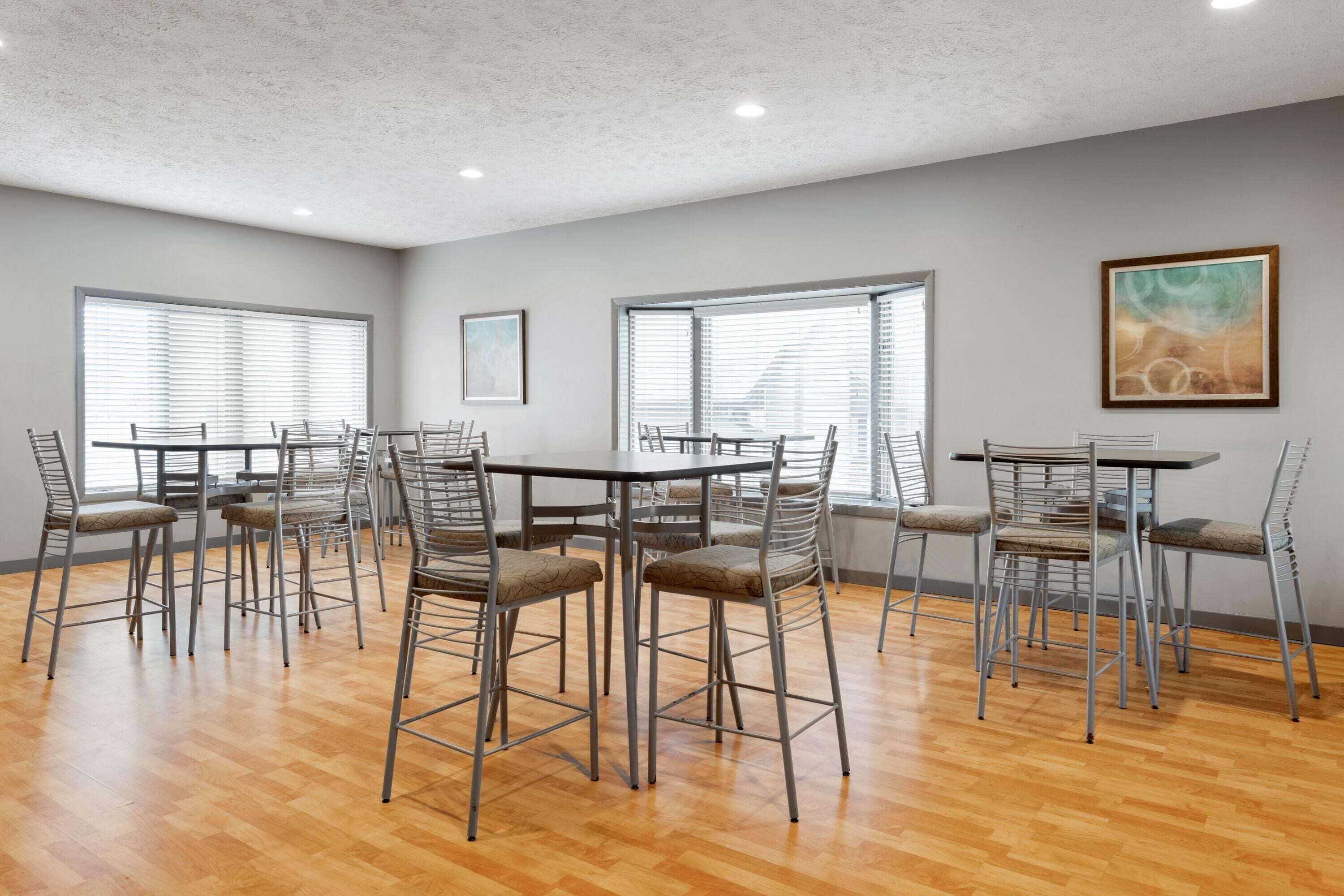 A spacious room with multiple tables and chairs, large windows with blinds, and a ceiling with recessed lighting.