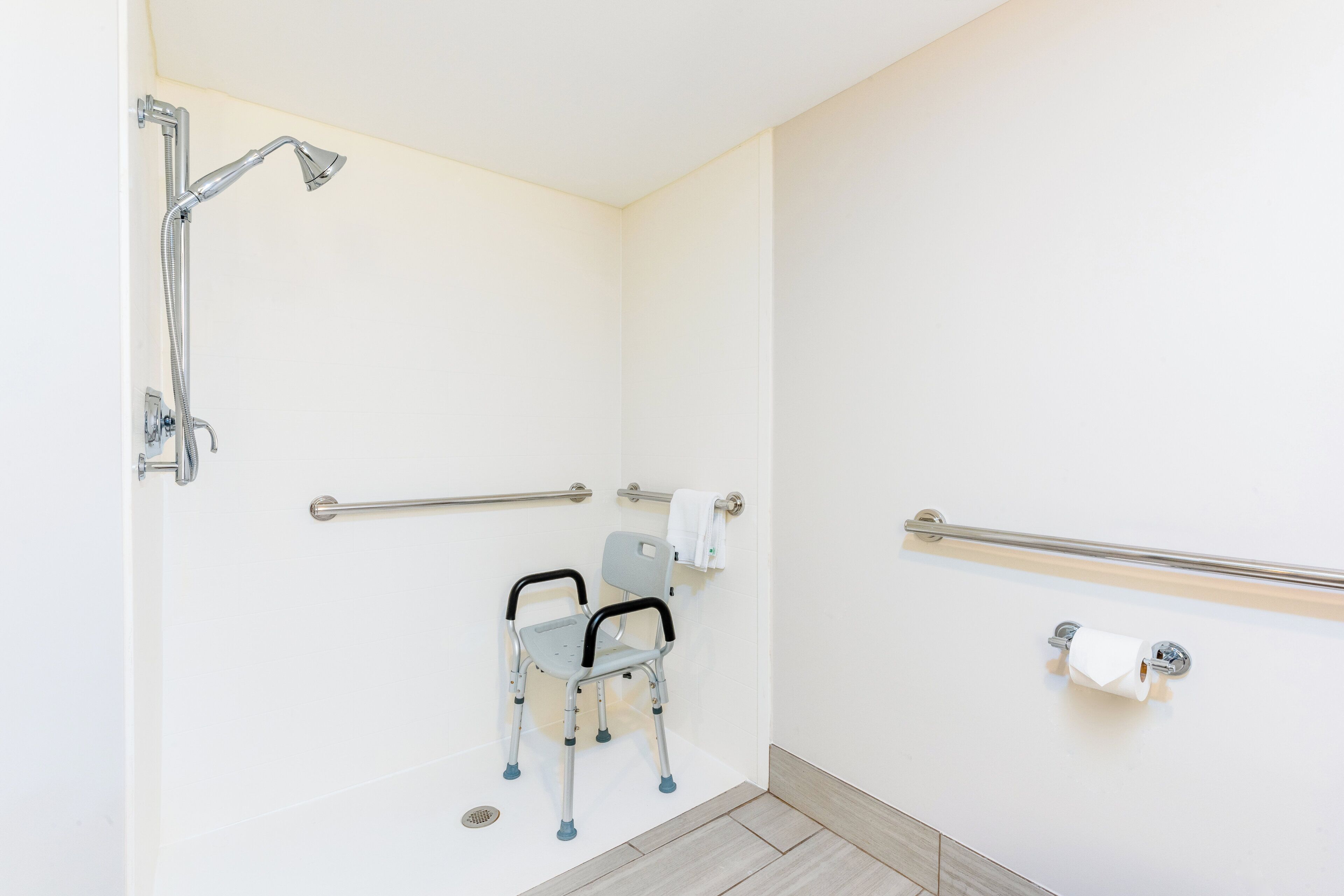 A wheelchair-accessible shower area with grab bars, a toilet with a paper towel holder, and a handrail.