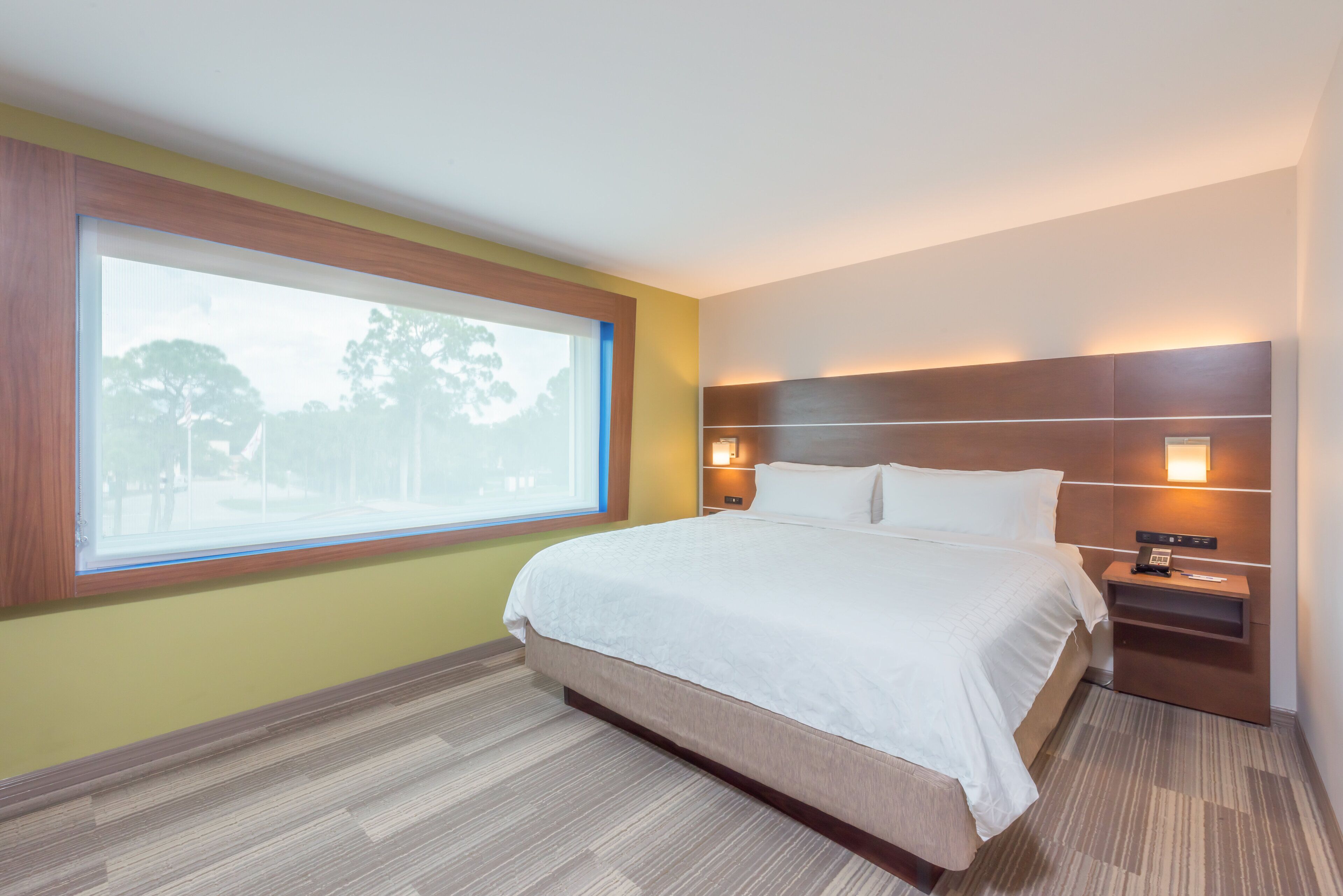 Habitación de hotel con un ventanal grande, una cama, una mesita de noche y un aplique de pared.
