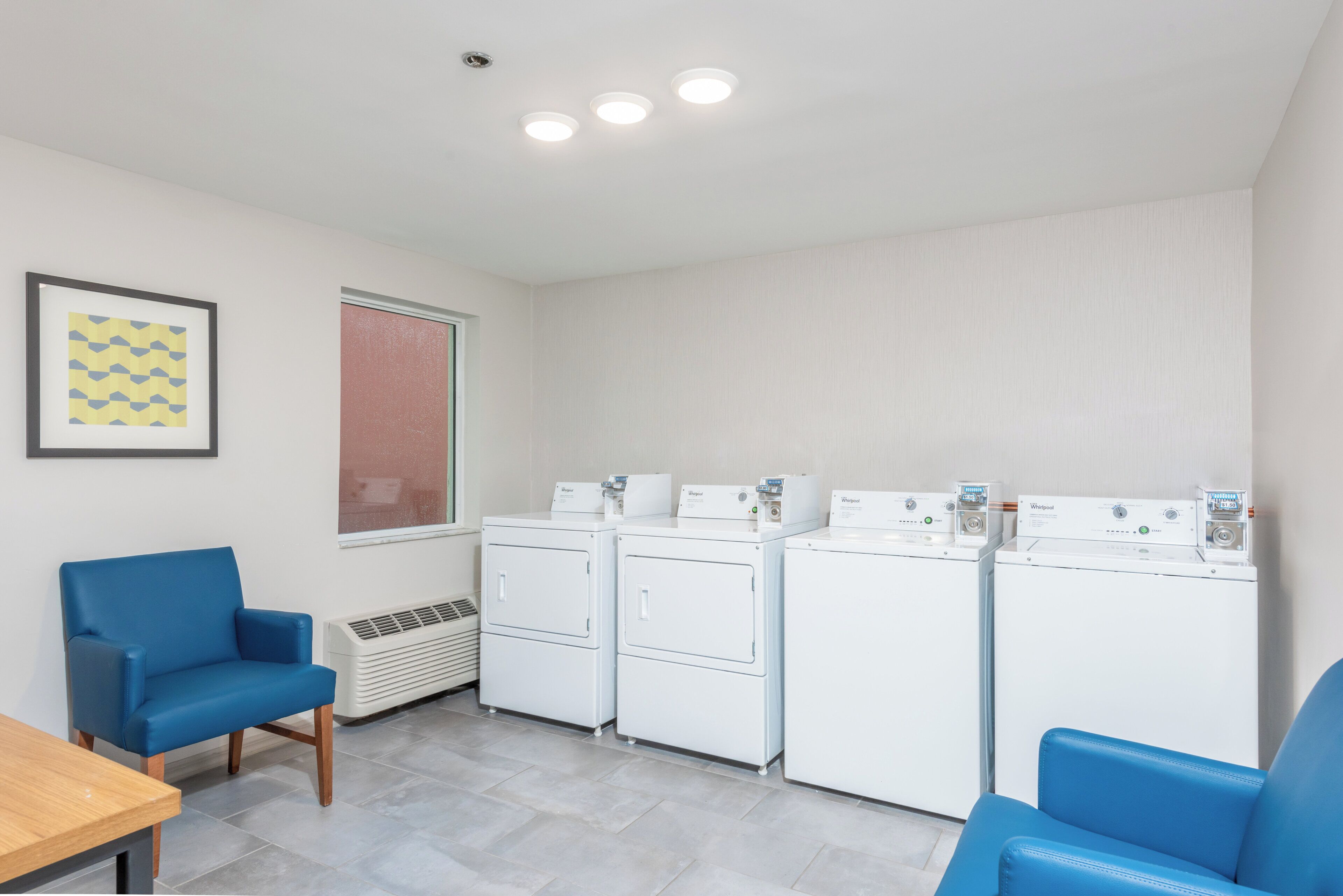 Un cuarto de lavandería con cuatro lavadoras y secadoras Whirlpool blancas, una silla azul, una mesa y un cuadro en la pared.
