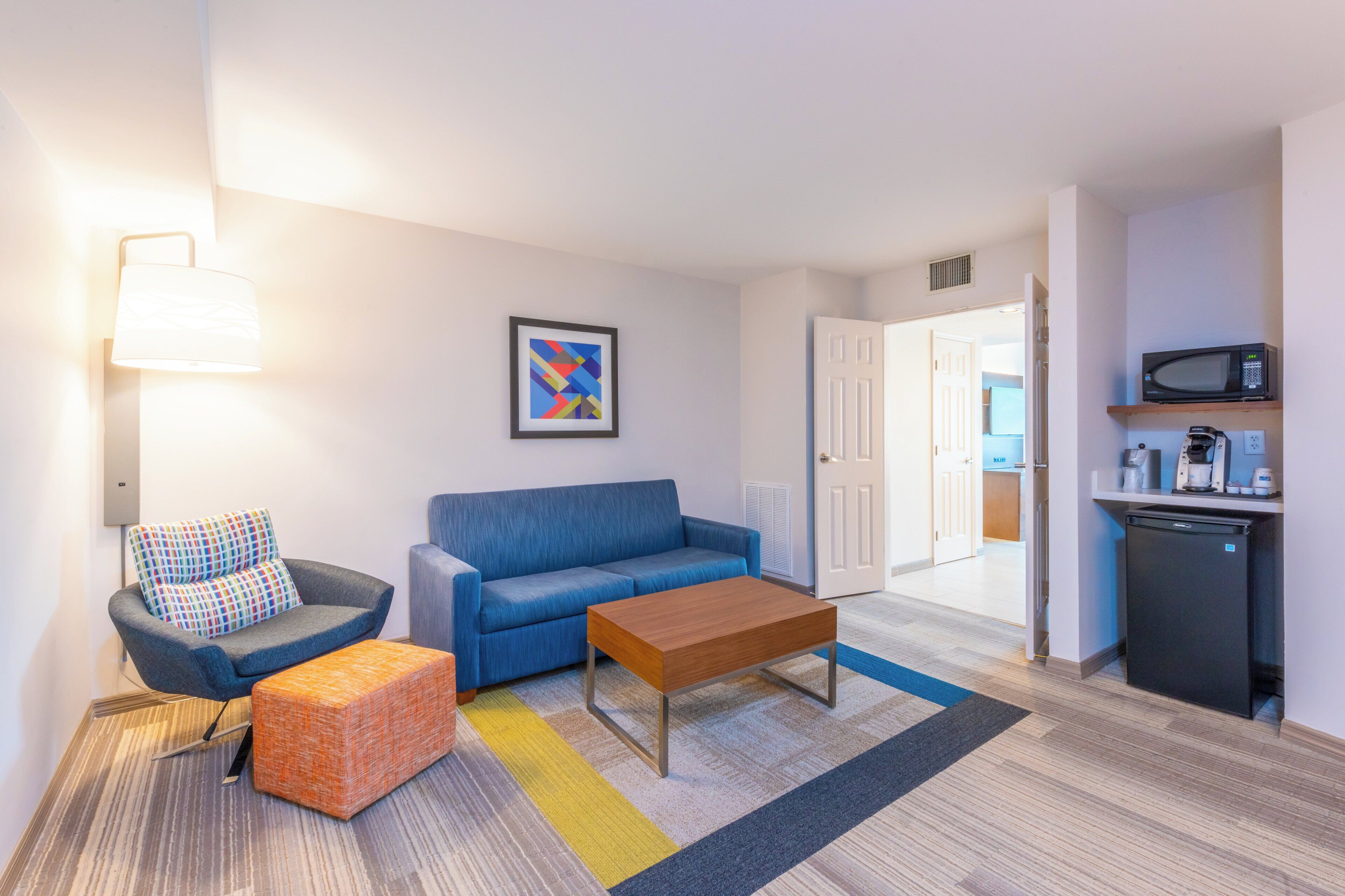 A compact living space with a sofa, a coffee table, a lamp, a colorful rug, and a kitchen area with a microwave and refrigerator.