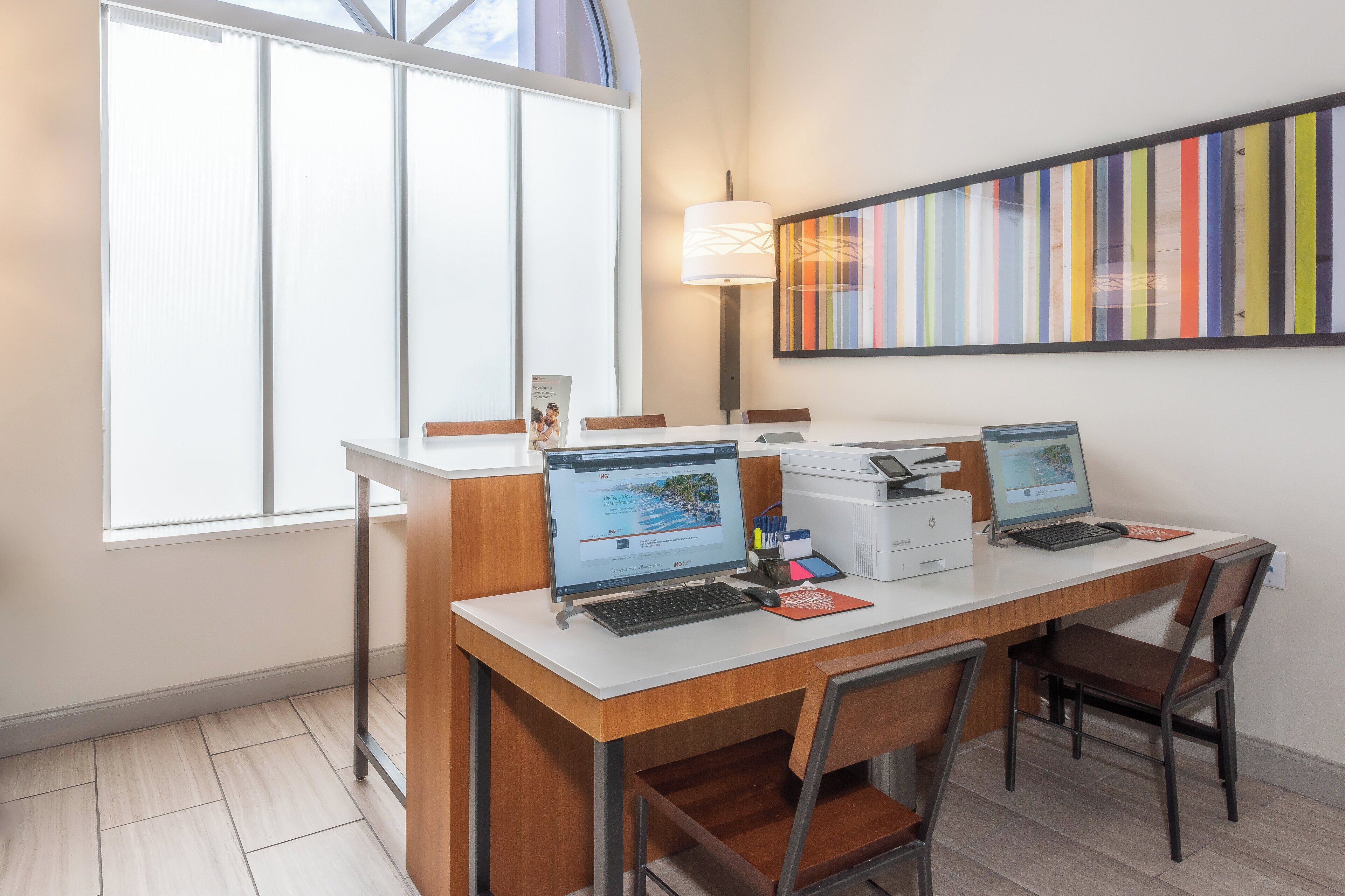 A modern office space with two desks, each with a computer, a printer, and a lamp. There is a colorful striped artwork on the wall and a window providing natural light.