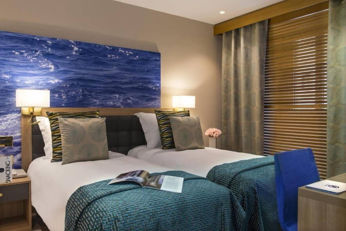 A hotel room with a bed, two bedside lamps, a large ocean-themed wall mural, and a window with blinds.