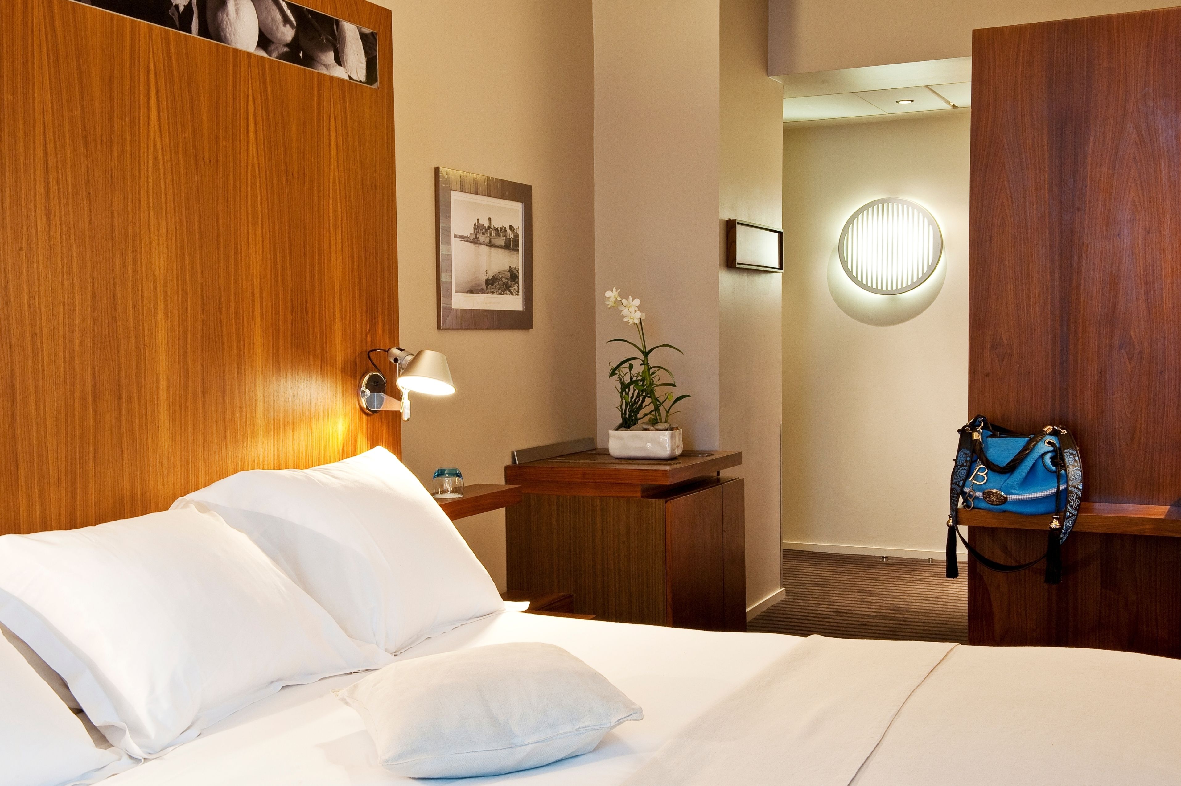 A hotel room with a wooden headboard, a bed with white linens, a nightstand with a lamp, a framed photo on the wall, a small table with a potted plant, and a blue bag hanging on a wooden shelf.