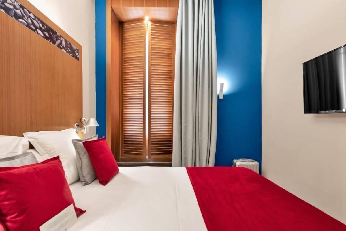 A hotel room with a bed, pillows, a nightstand, a lamp, a TV, and a window with curtains.