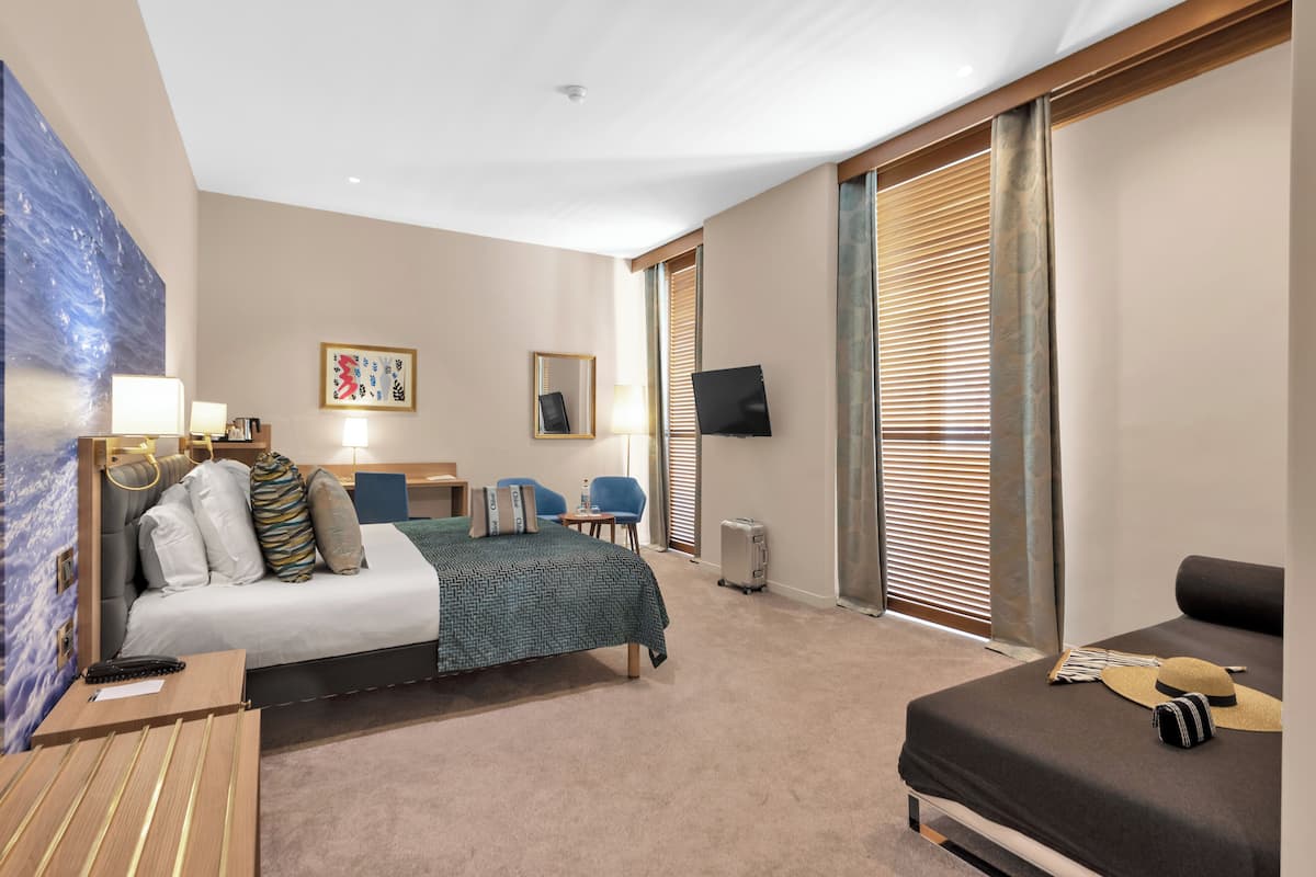 A hotel room with a bed, a desk, a chair, a TV, and a large window with blinds.