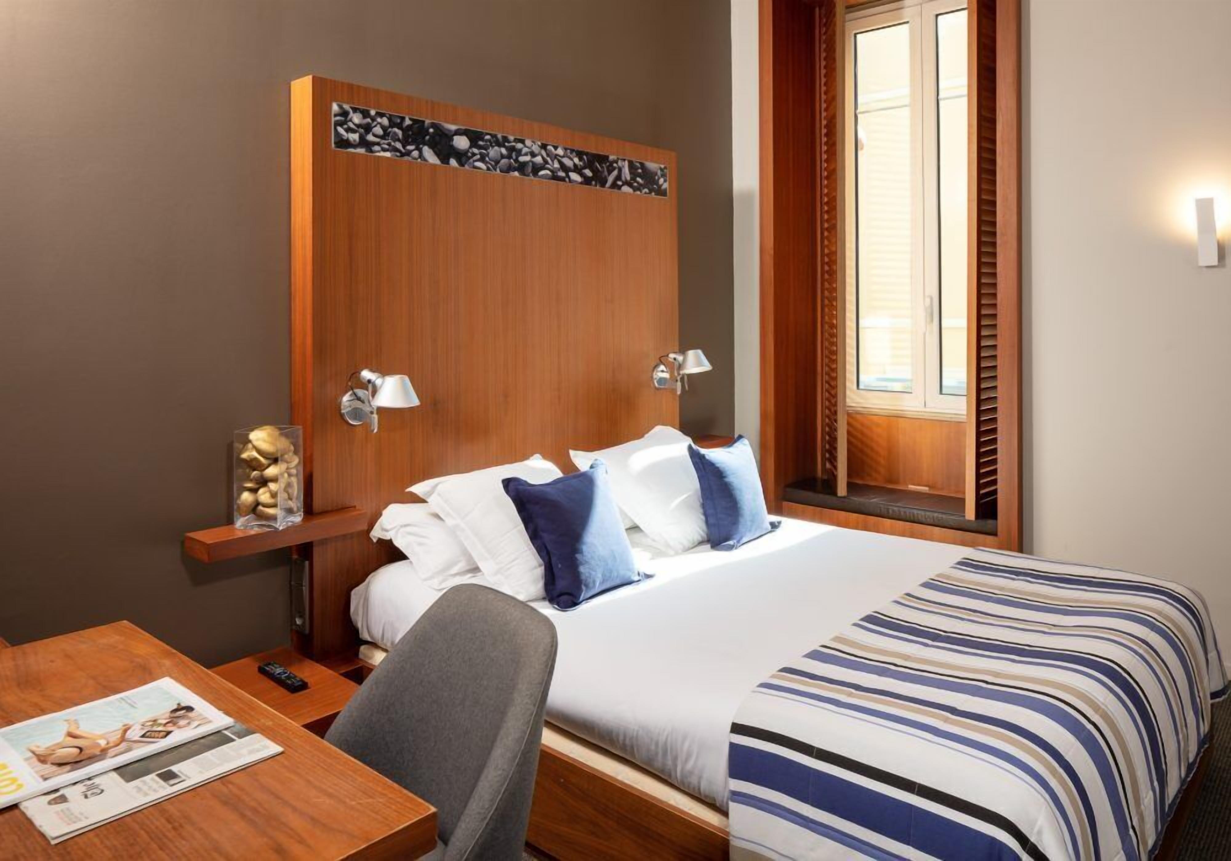 Une chambre d’hôtel avec une tête de lit en bois, un lit avec une literie à rayures, une table de chevet avec une lampe, une chaise et un magazine posé sur la table.