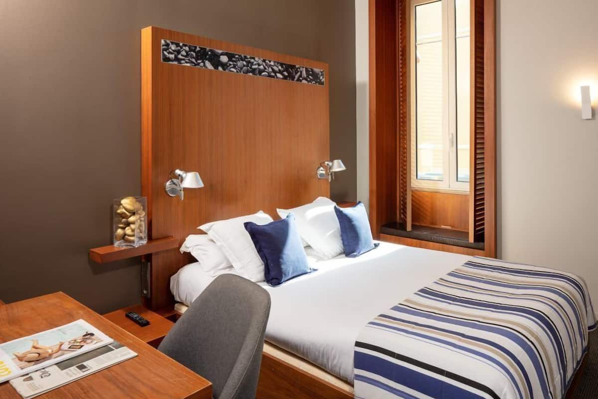 Ein Hotelzimmer mit Holz-Kopfteil, einem Bett mit gestreifter Bettwäsche, einem Nachttisch mit Lampe, einem Sessel und einer Zeitschrift auf dem Tisch.