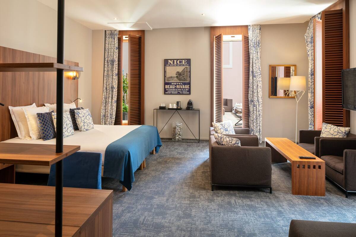 A hotel room with a bed, a sofa, a desk, and a TV. There is a poster on the wall and a window with curtains.