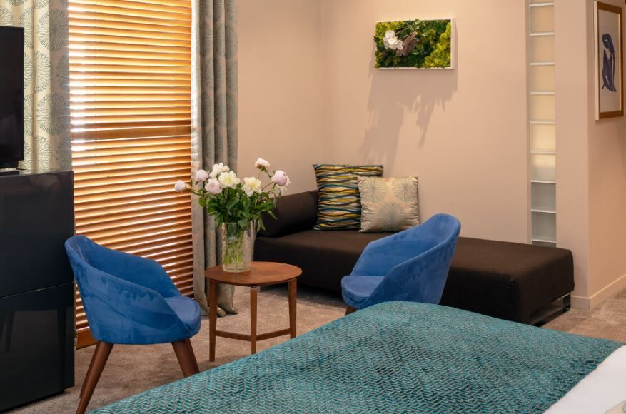 A modern living room with a sofa, two blue chairs, a small table with a vase of flowers, a television, and a painting on the wall.